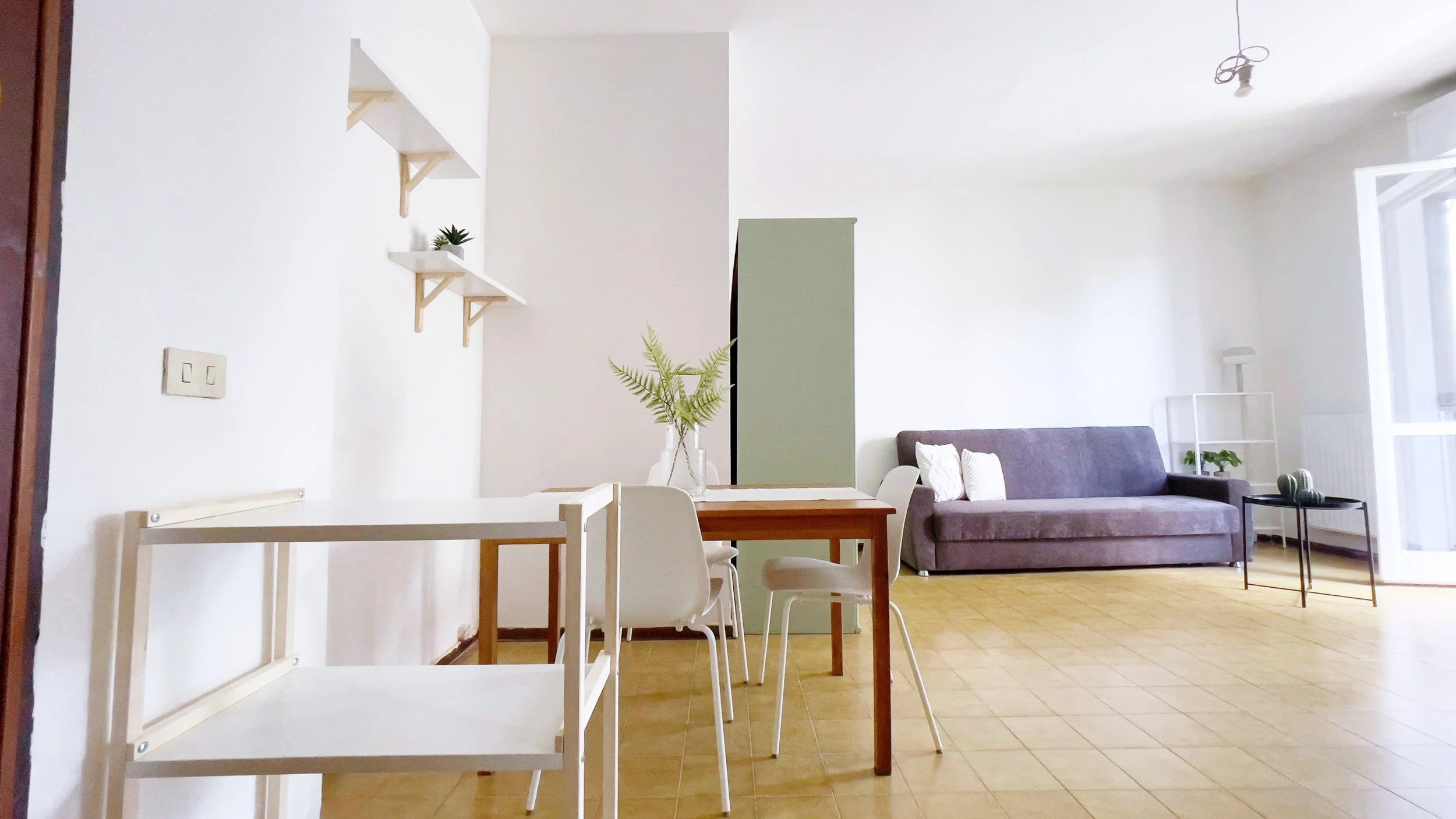 Interno di un soggiorno moderno con divano grigio, tavolo da pranzo in legno e sedie bianche, scaffali a muro con piante decorative, pavimento in piastrelle.