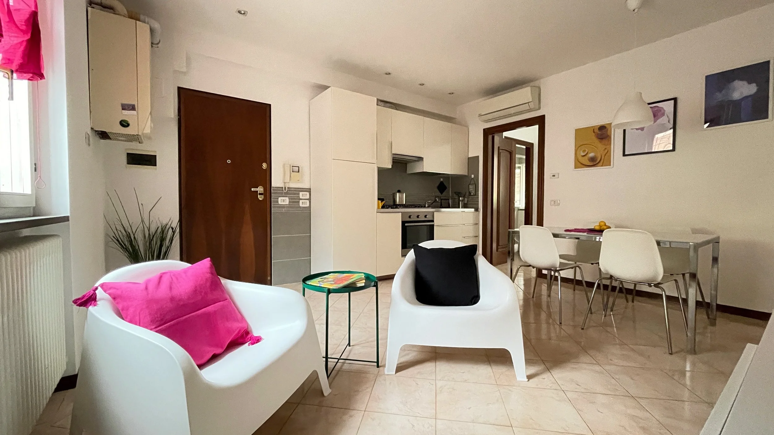 Interno di un appartamento moderno con salotto e cucina a vista. Due sedie bianche con cuscini colorati, un tavolino verde, cucina bianca con forno, tavolo da pranzo con sedie bianche, pavimento in piastrelle beige e opere d'arte appese al muro.