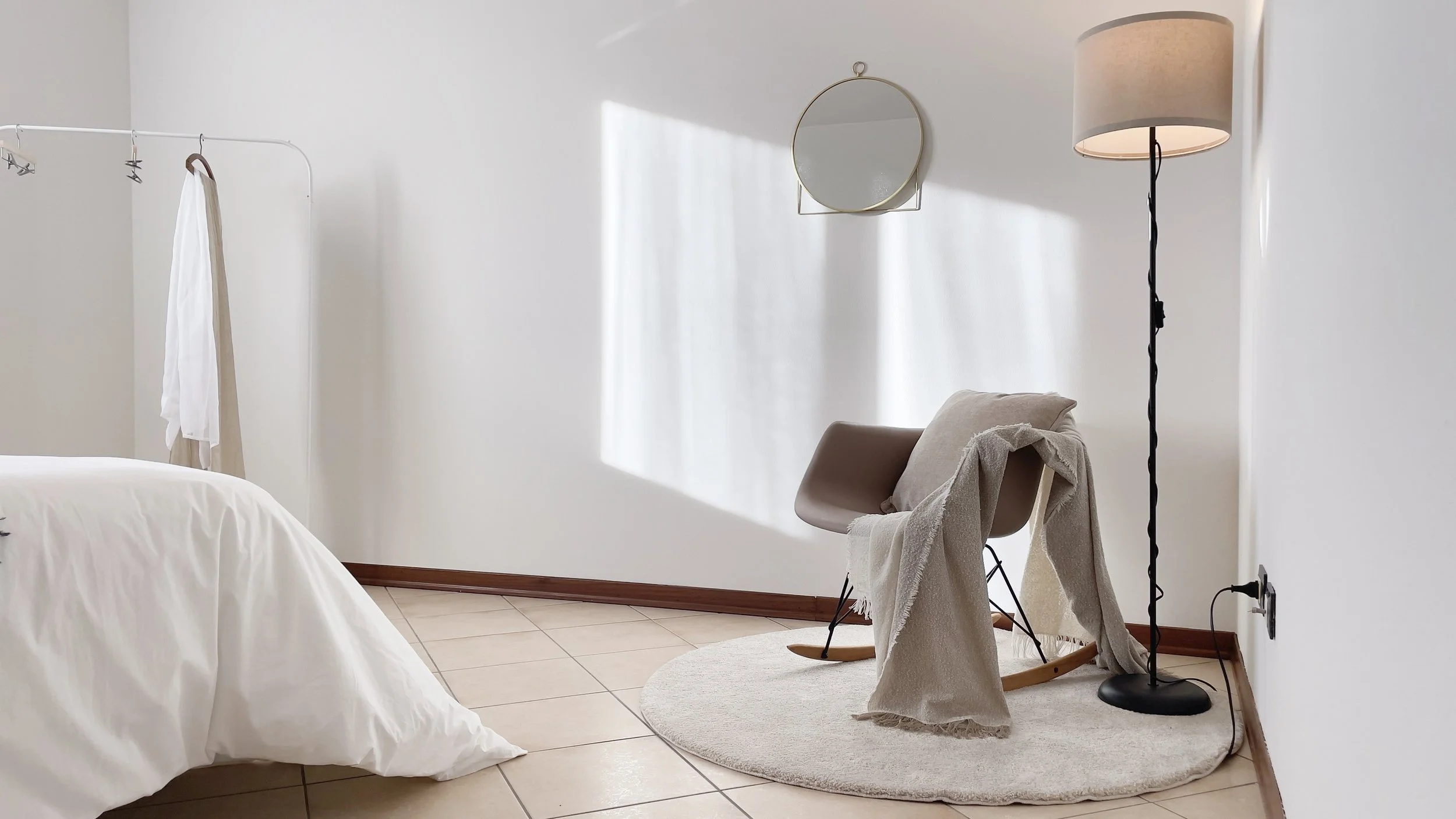 Camera da letto minimalista con letto bianco, sedia con coperta, lampada da terra, specchio rotondo e tappeto chiaro.