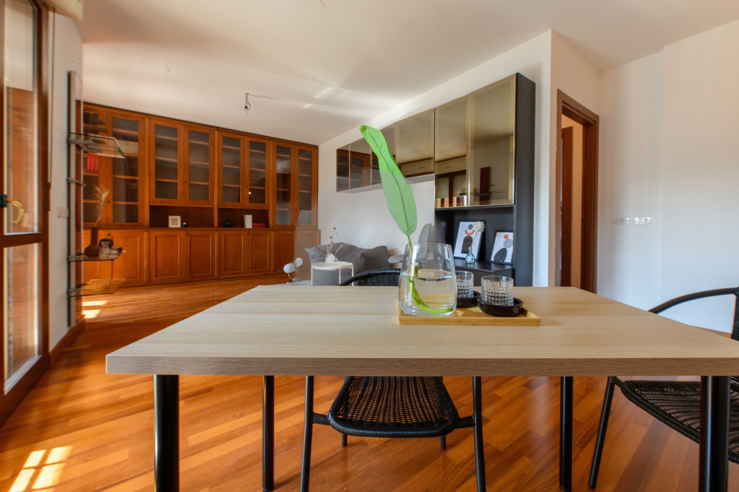 Sala da pranzo moderna con tavolo in legno chiaro, sedia nera, vaso trasparente con foglia verde, pavimento in legno, scaffalature in legno scuro e mobili moderni.
