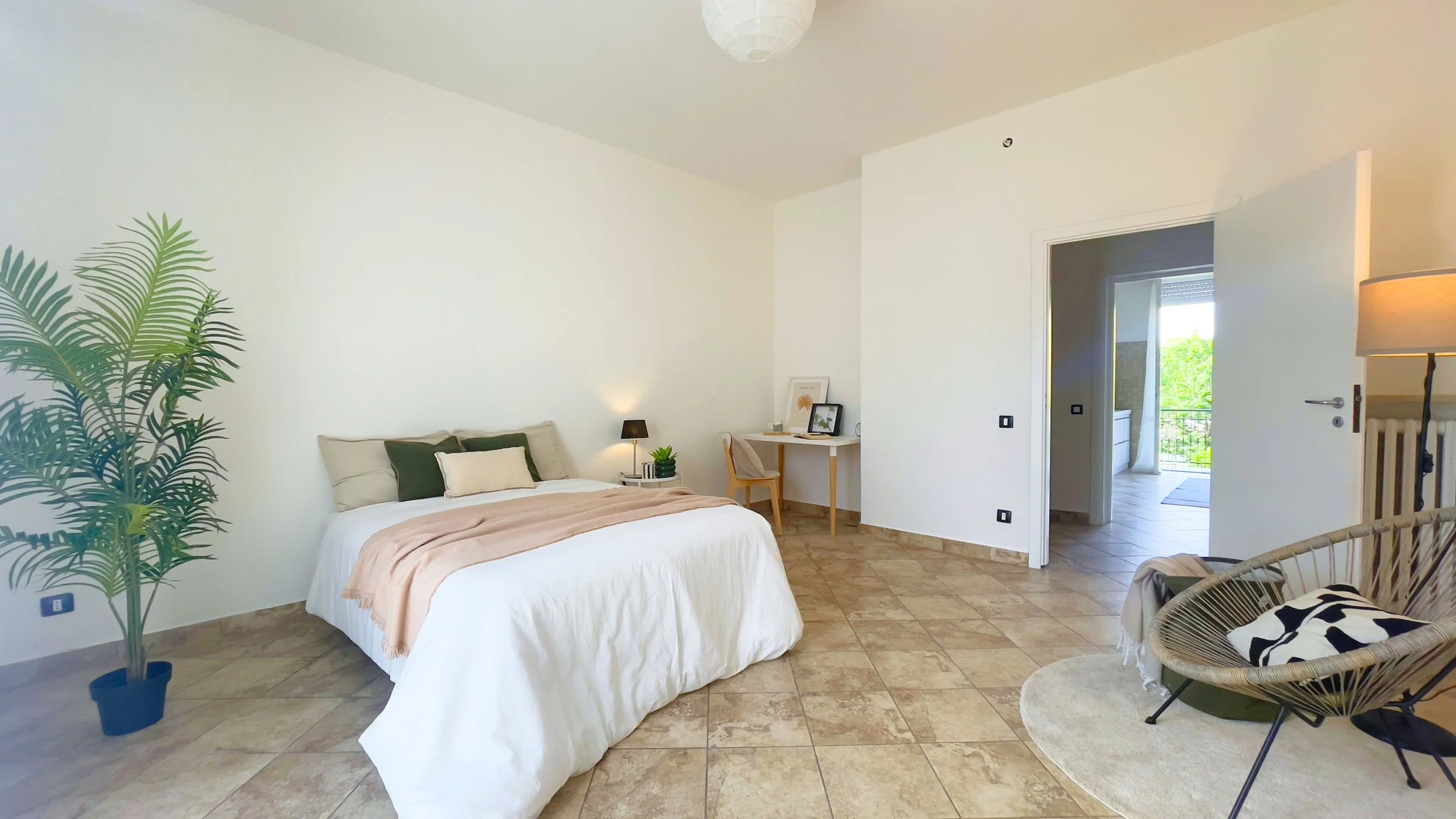 Camera da letto minimalista con letto matrimoniale, pianta decorativa, scrivania con sedia, poltrona e porta aperta verso il balcone.