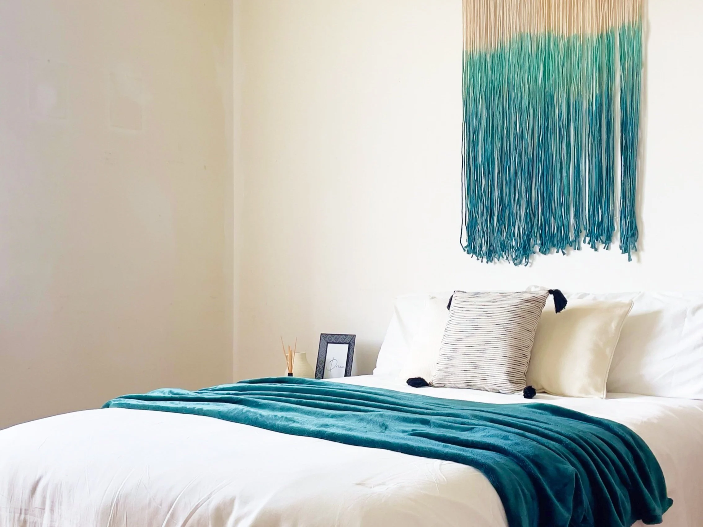 Interno di una camera da letto con letto matrimoniale, coperta blu petrolio, cuscini bianchi e decorazione murale in filato turchese.