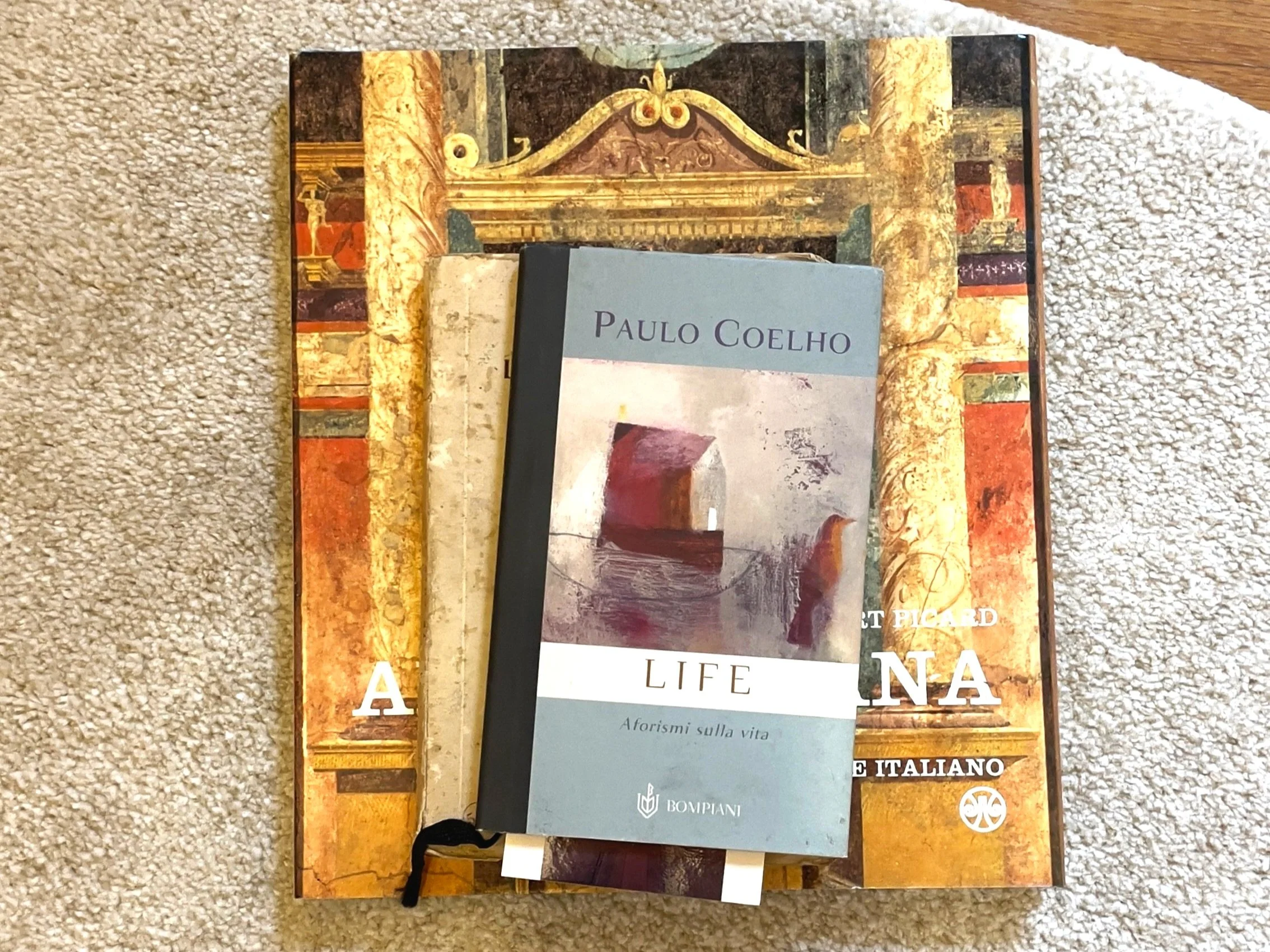 Libri impilati su un tappeto, con un libro di Paulo Coelho in cima. I libri sotto hanno copertine con motivi classici.