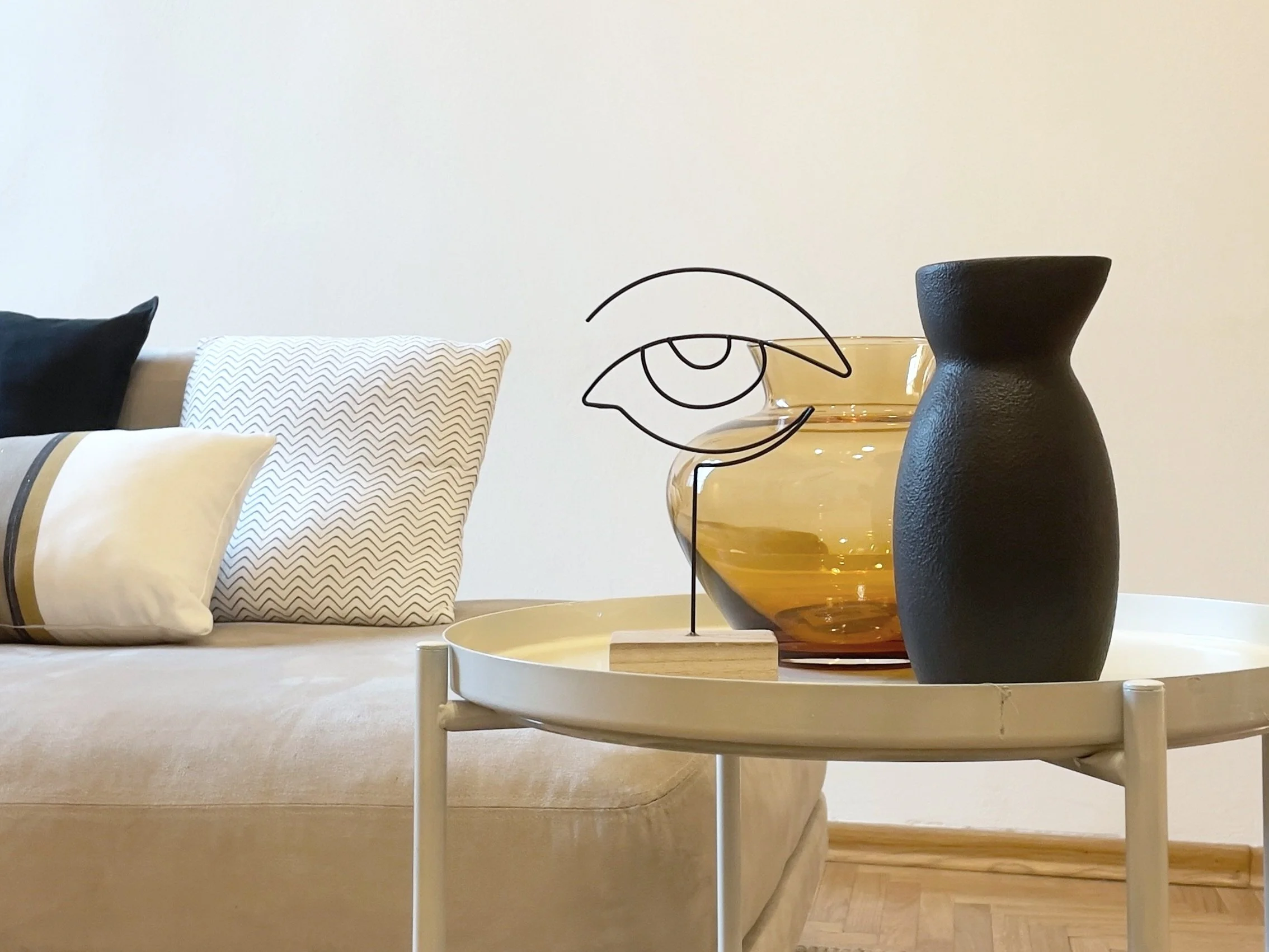 Soggiorno con divano beige, cuscini bianchi e neri, tavolino rotondo con vaso nero e decorazione artistica, vaso di vetro ambrato sullo sfondo.