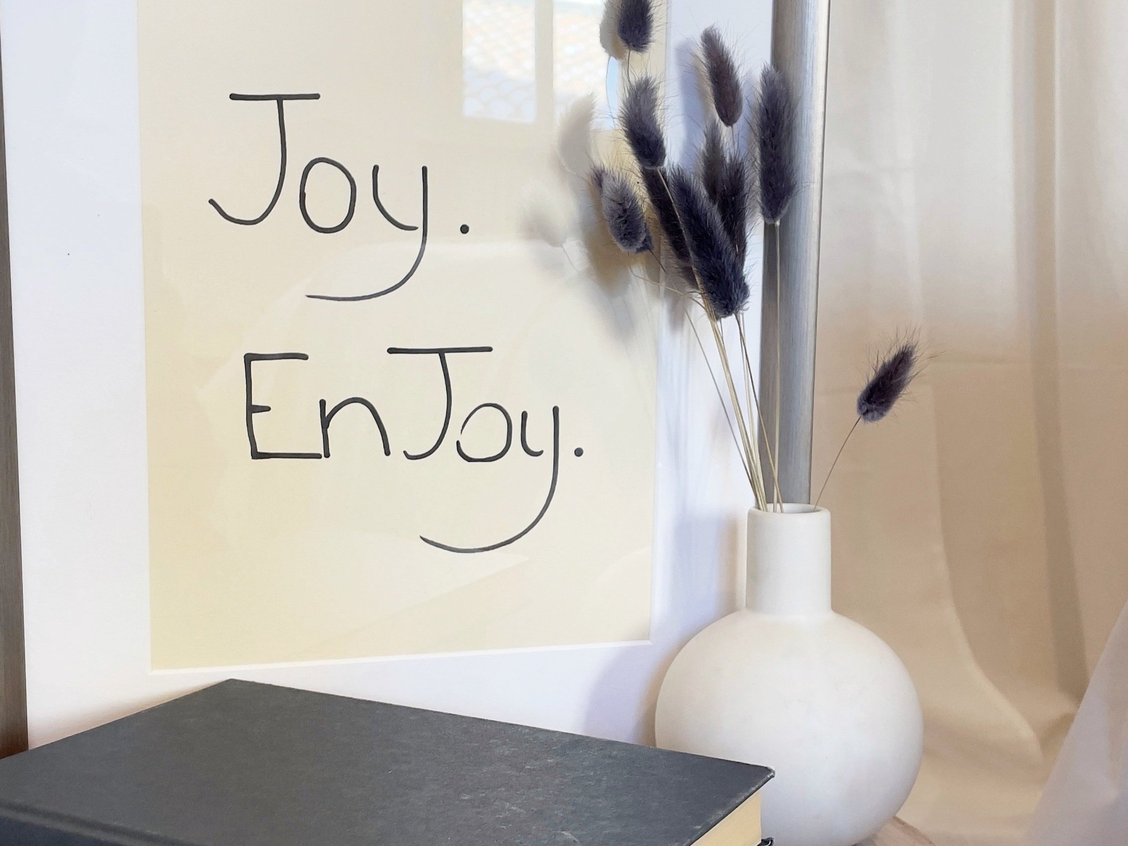 Cornice con testo 'Joy. EnJoy.' accanto a vaso bianco con piume.