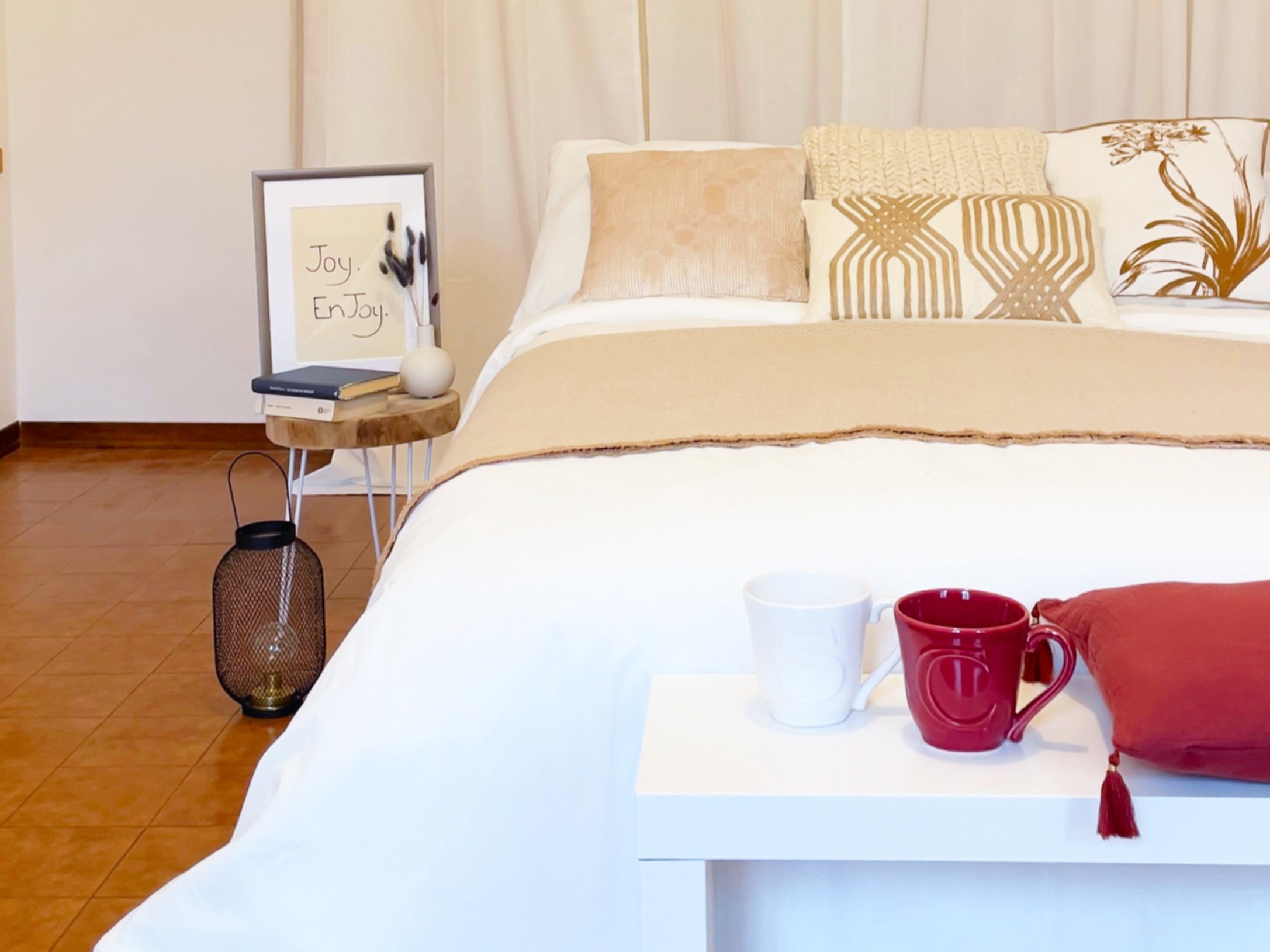 Camera da letto moderna con letto matrimoniale e cuscini decorativi. Un tavolino laterale con libri, un vaso e una cornice con la scritta "Joy. EnJoy.". Lume a forma di lanterna sul pavimento. Due tazze e un cuscino rosso su un tavolino bianco ai pie