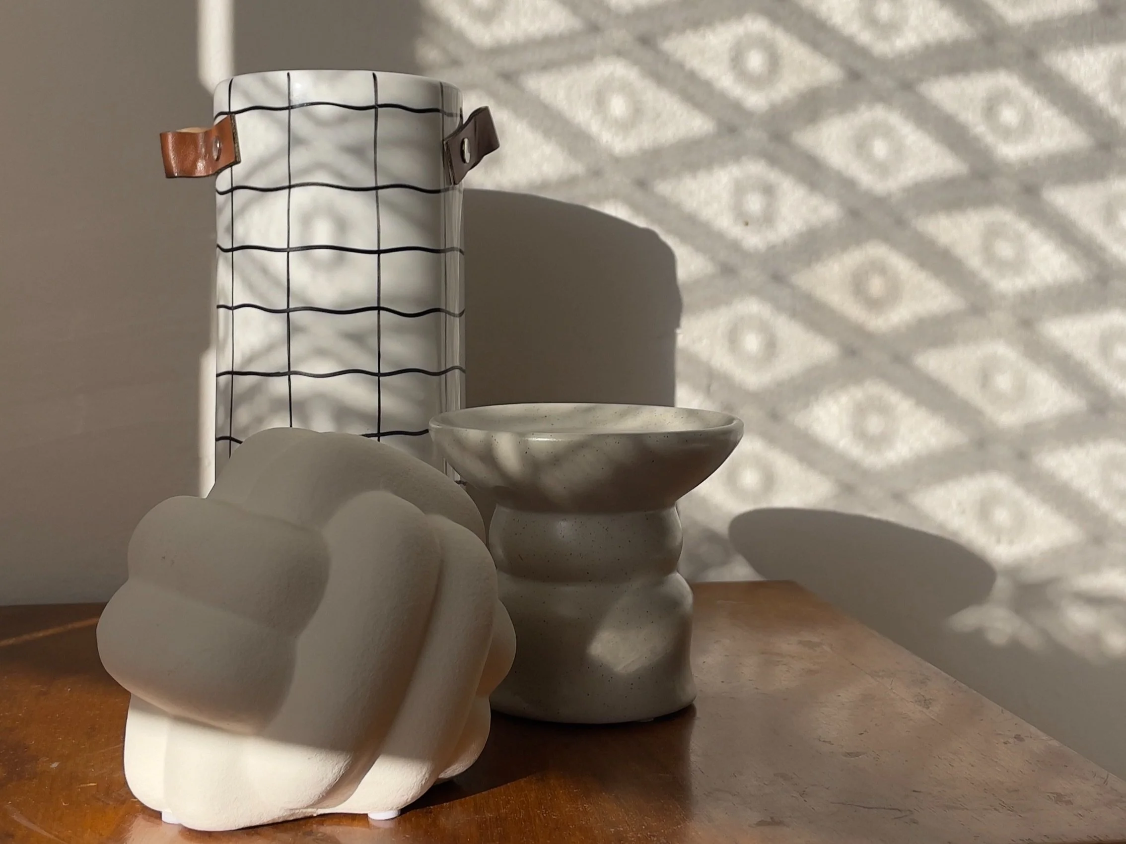 Immagine di un vaso bianco con motivi a griglia, una scultura bianca astratta e un oggetto cilindrico su un tavolo di legno, con ombre decorative sullo sfondo.