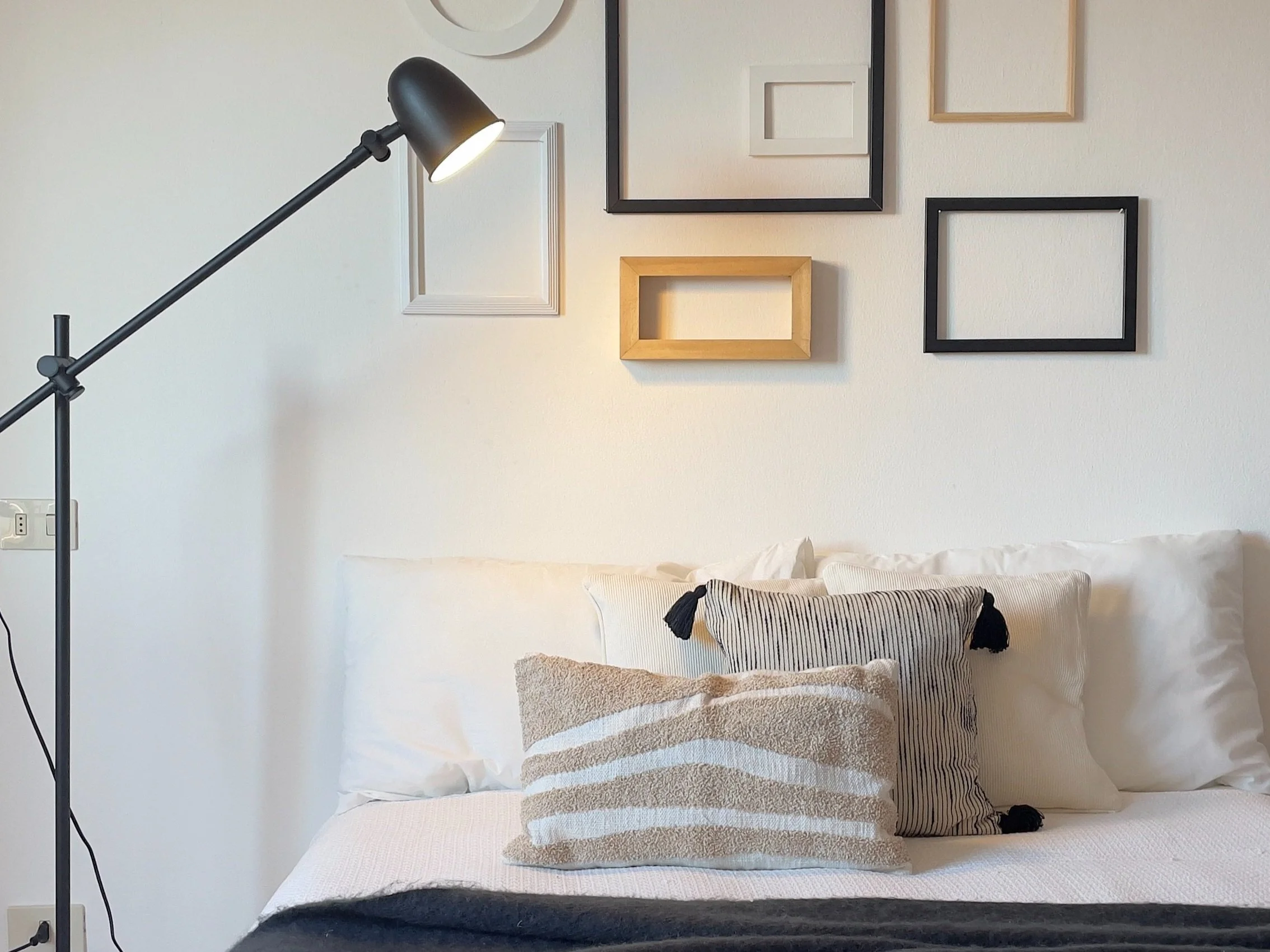 Interno di una camera da letto con letto matrimoniale, cuscini decorativi, lampada da terra nera e cornici appese al muro.