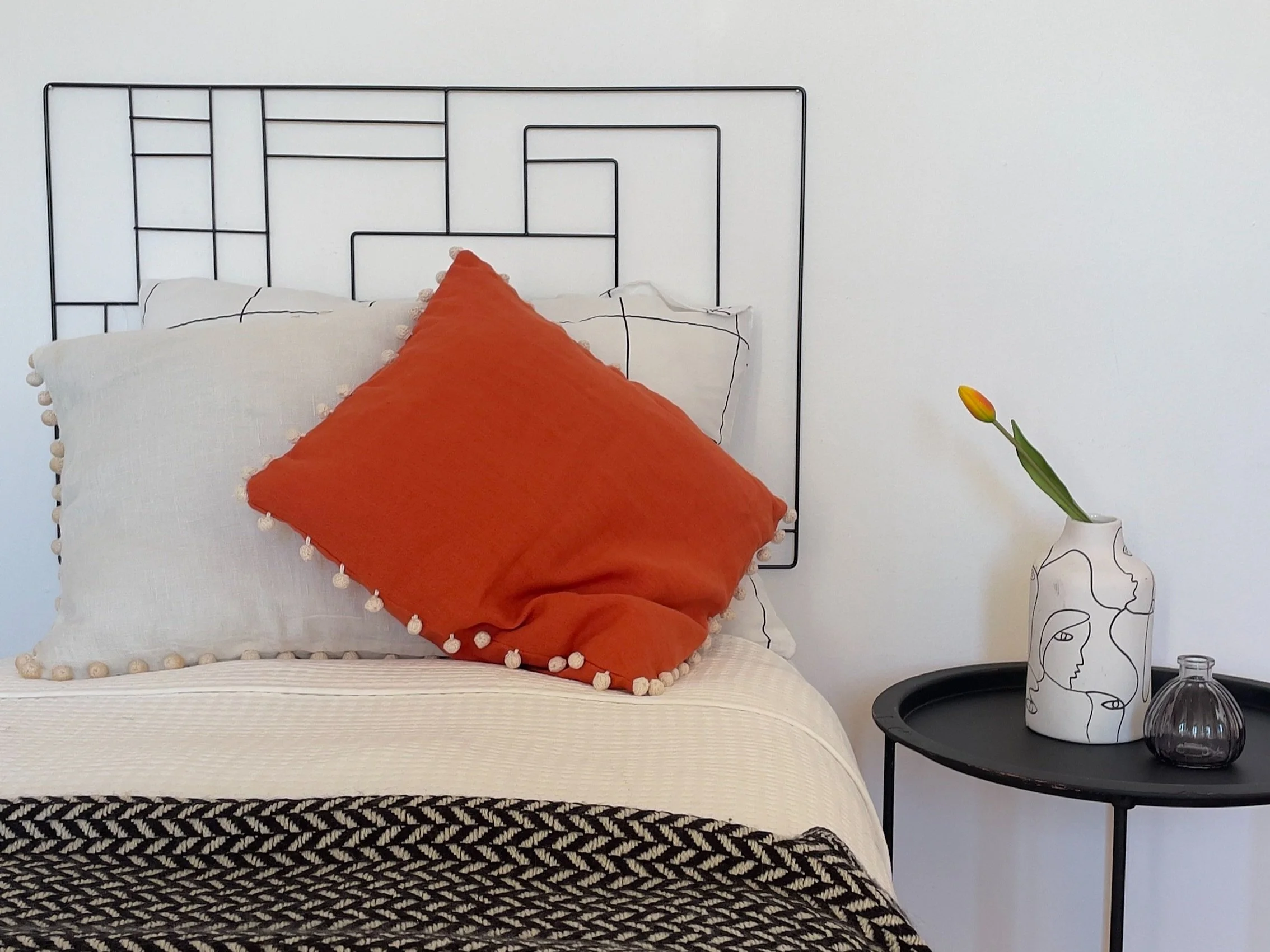 Letto con cuscini e coperta decorativa, testiera geometrica in metallo nero. A destra, un vassoio rotondo nero con un vaso bianco contenente un tulipano giallo.