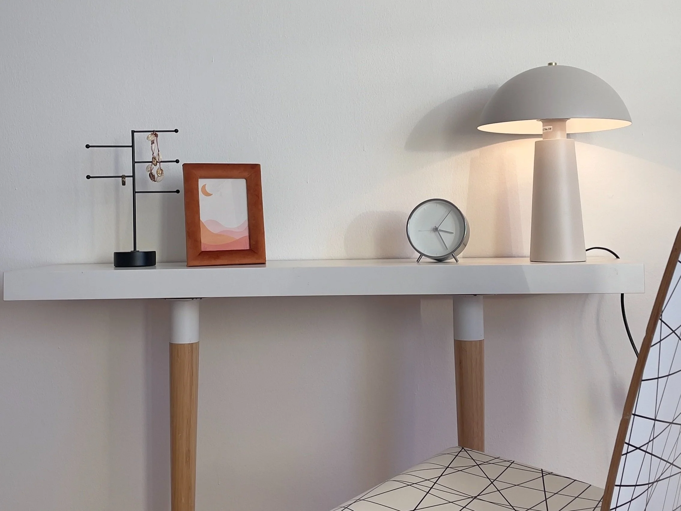 Mensola con lampada da tavolo, orologio, portagioie, cornice con immagine astratta, e sedia parzialmente visibile.
