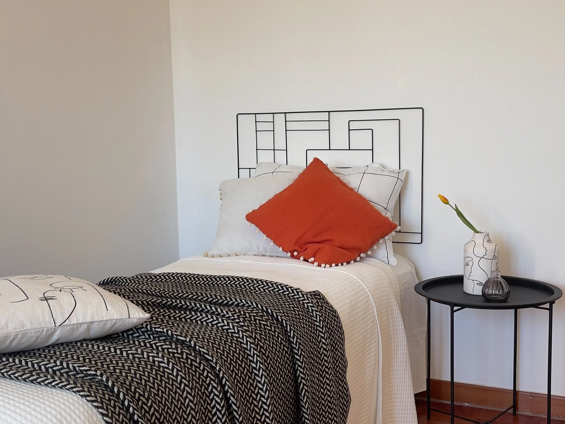 Camera da letto moderna con letto singolo, cuscino arancione, copriletto a motivi geometrici, tavolino nero a lato, vaso decorativo con fiore giallo