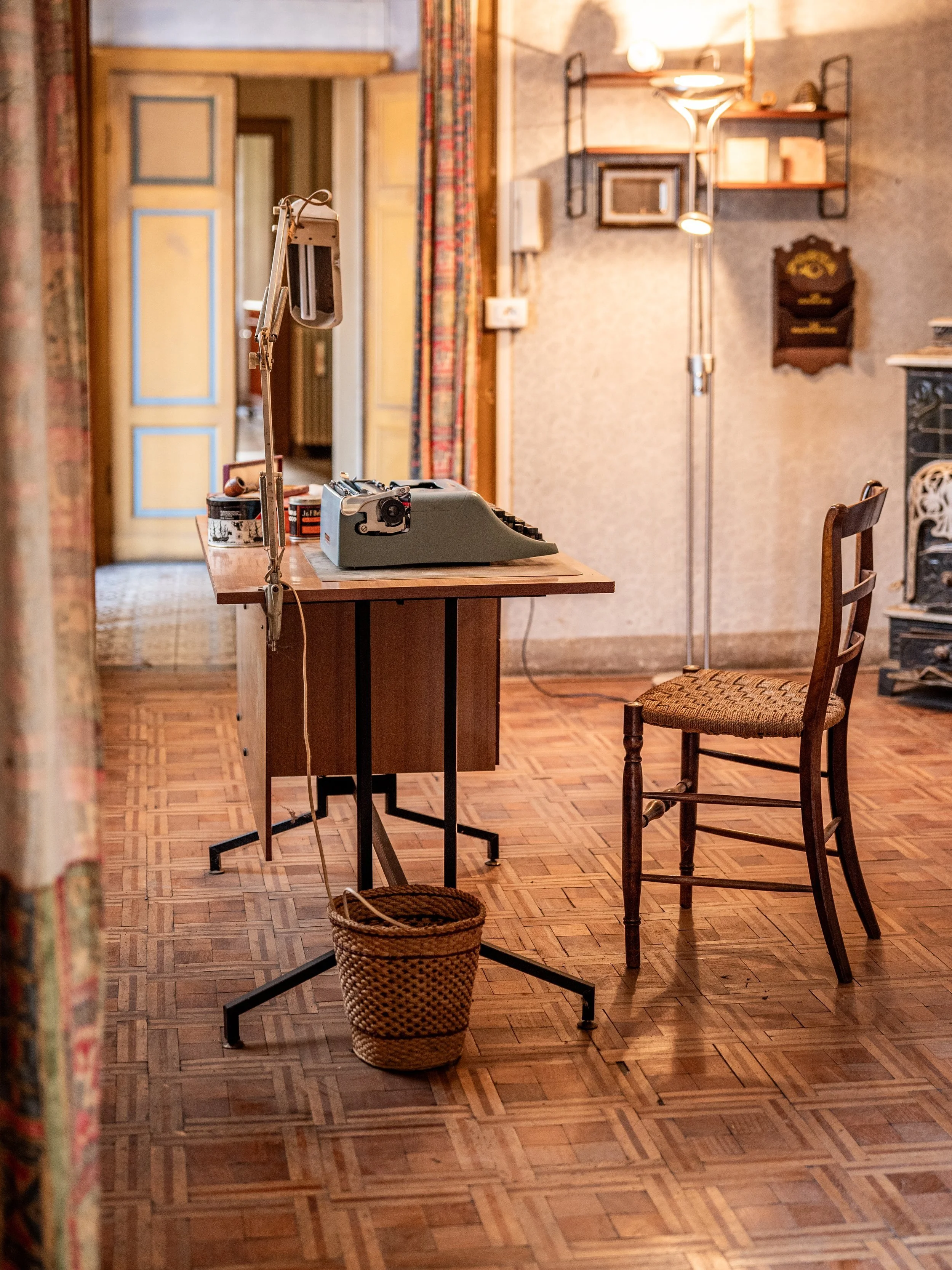 Interno di una stanza con una macchina da scrivere su un tavolo, una sedia di legno, un cestino intrecciato e un lampadario vintage. Sullo sfondo si vede una porta aperta e uno scaffale con oggetti decorativi.