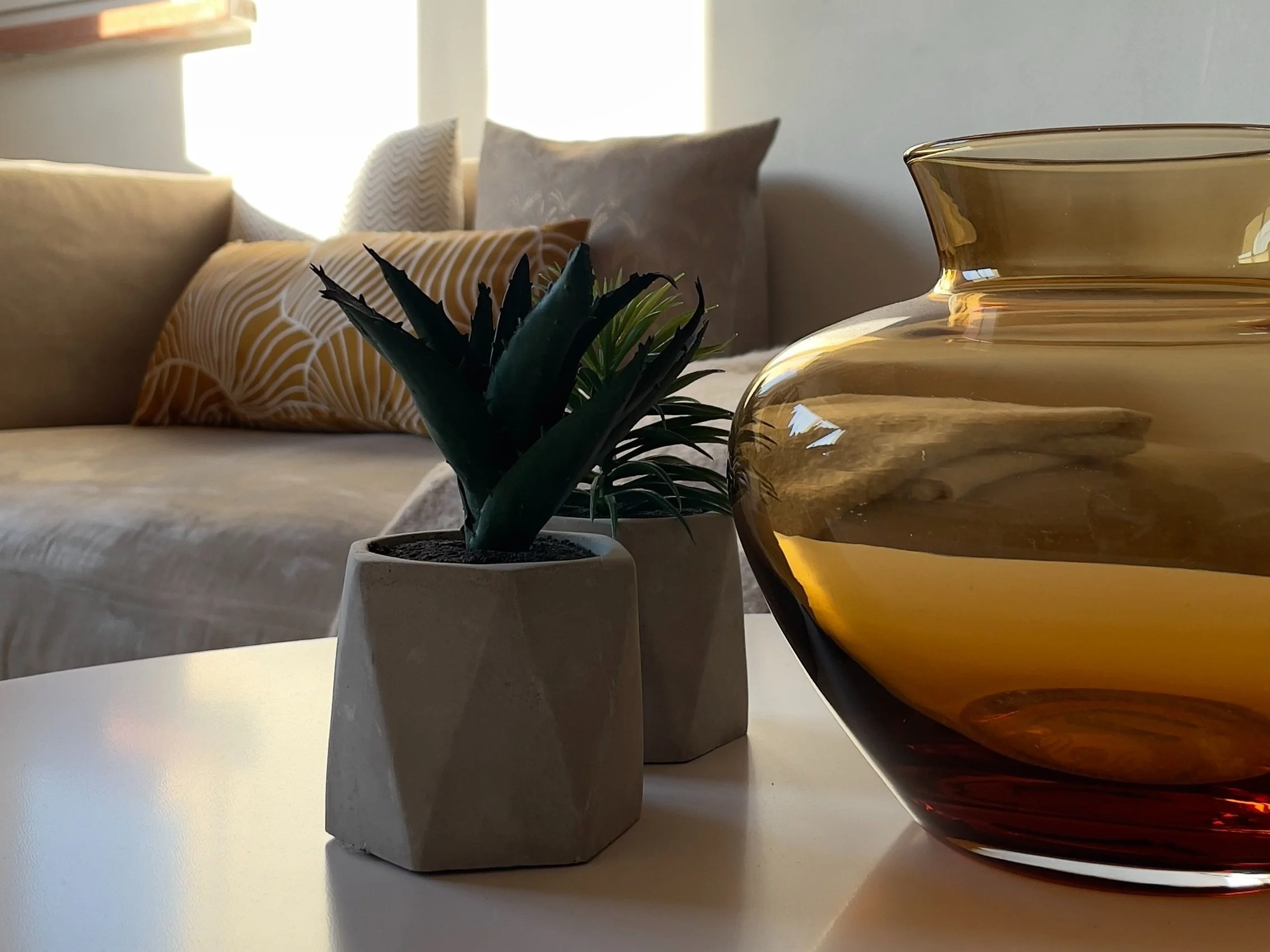 Interno di un soggiorno moderno con un divano beige, cuscini decorativi, un vaso di vetro marrone e una pianta in vaso su un tavolino bianco.