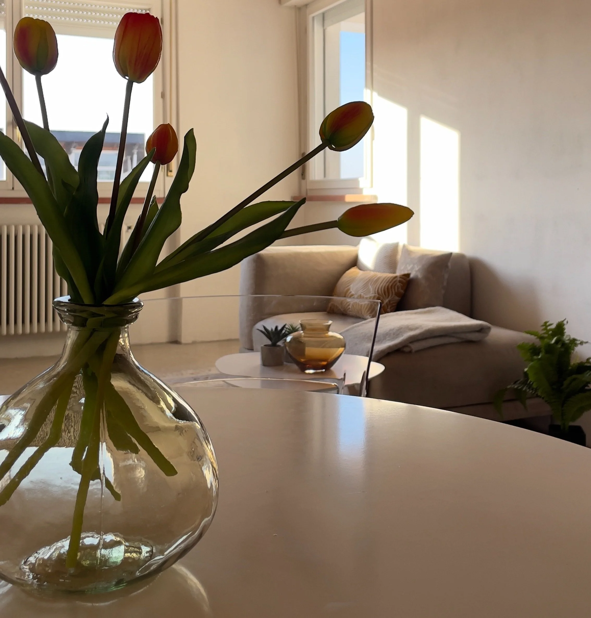 Interno di un salotto moderno con un vaso di tulipani su un tavolo e un divano beige sullo sfondo, illuminato dalla luce naturale.
