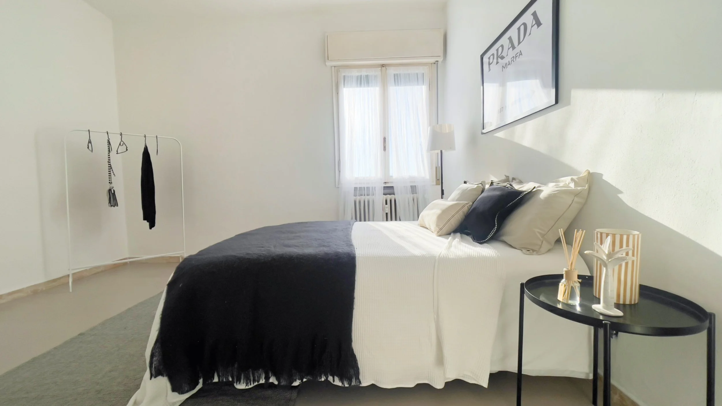 Camera da letto moderna con letto matrimoniale, biancheria in bianco e nero, appendiabiti minimalista, finestra con tende bianche e decorazioni semplici su tavolino.