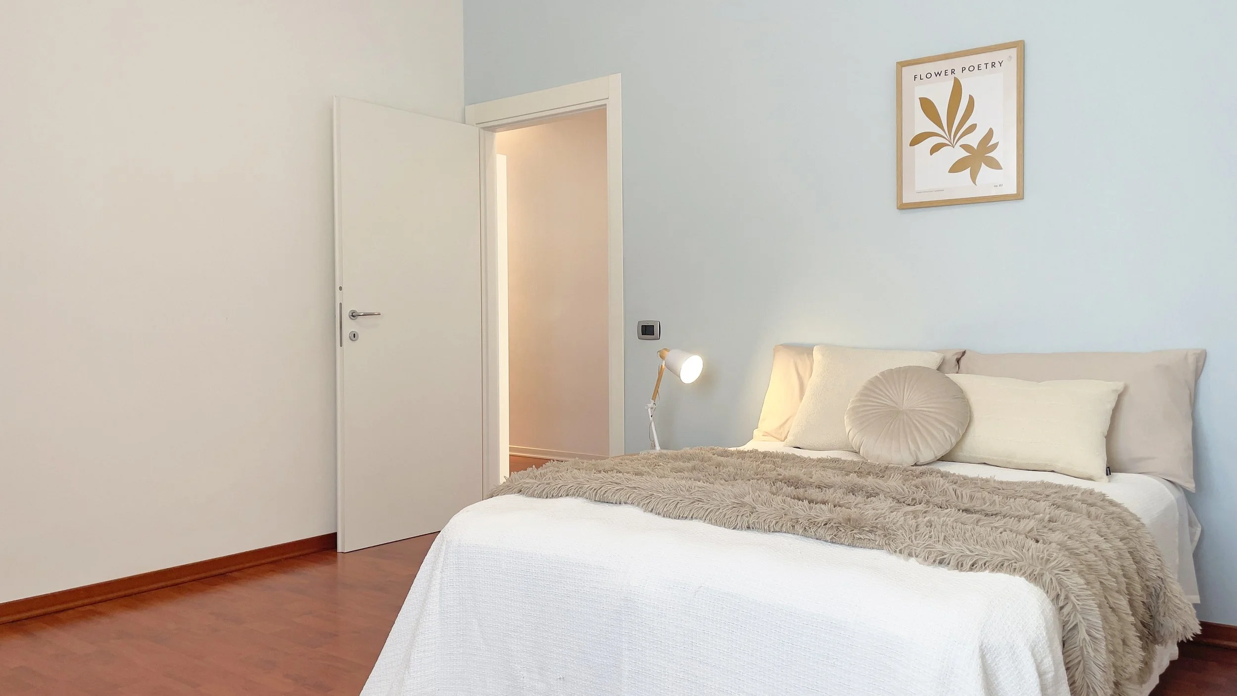 Camera da letto con letto coperto da coperte bianche e beige, lampada da tavolo bianca, parete con quadro e porta semiaperta.