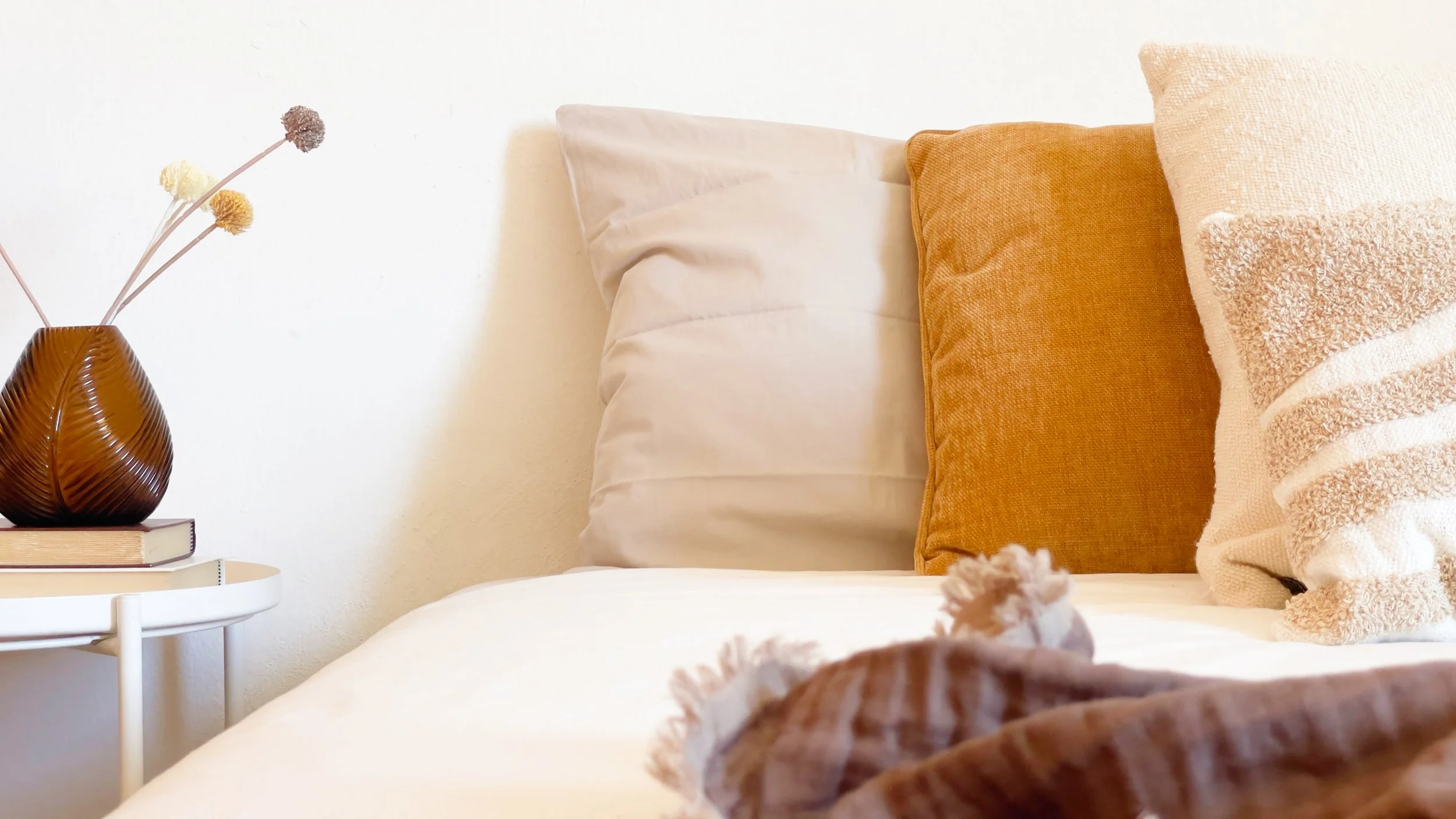 Interno di una camera da letto con cuscini colorati su un letto, un vaso marrone con decorazioni floreali secche su un comodino.