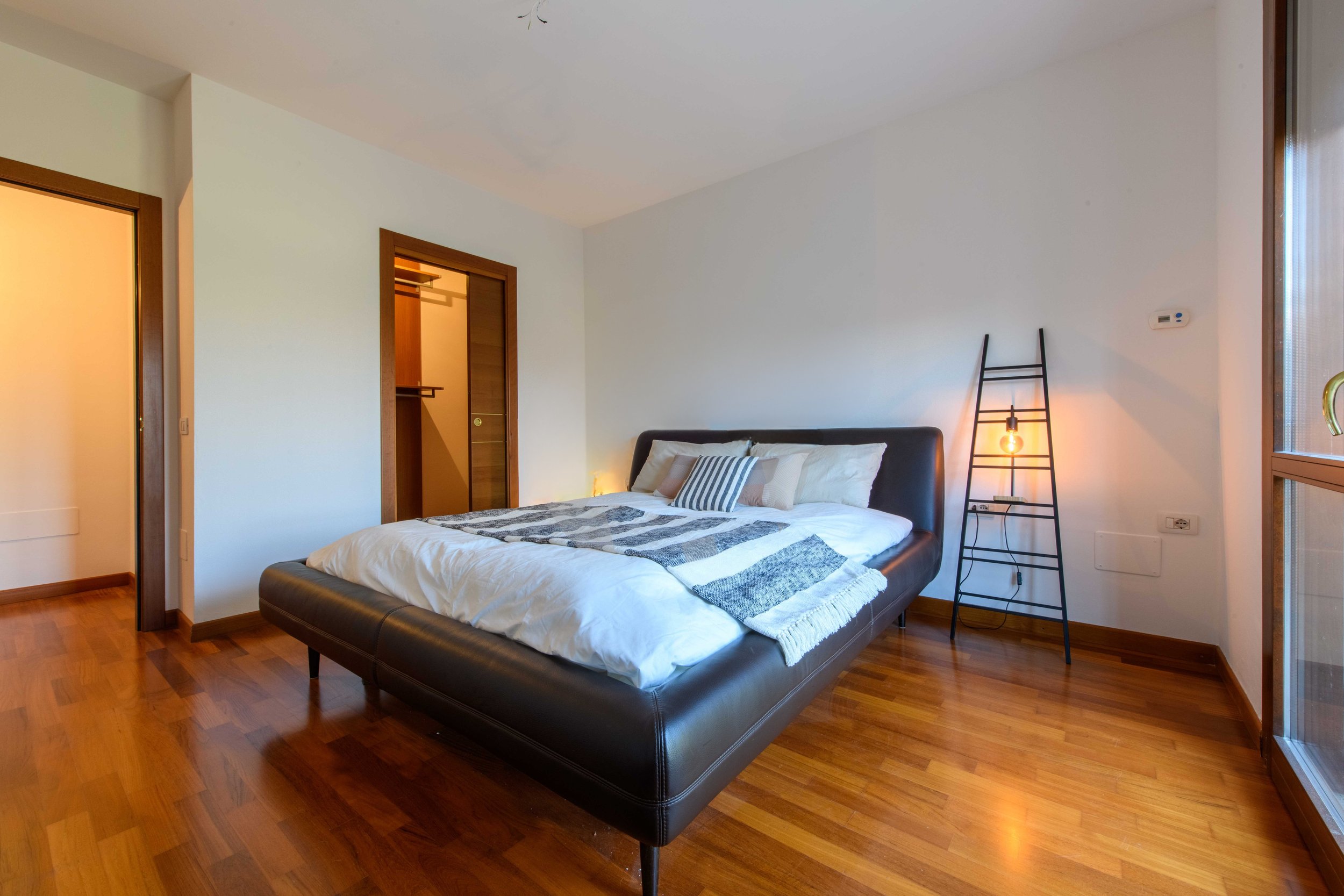 Camera da letto moderna con letto matrimoniale, pavimento in legno, lampada da terra e porta aperta verso una cabina armadio.