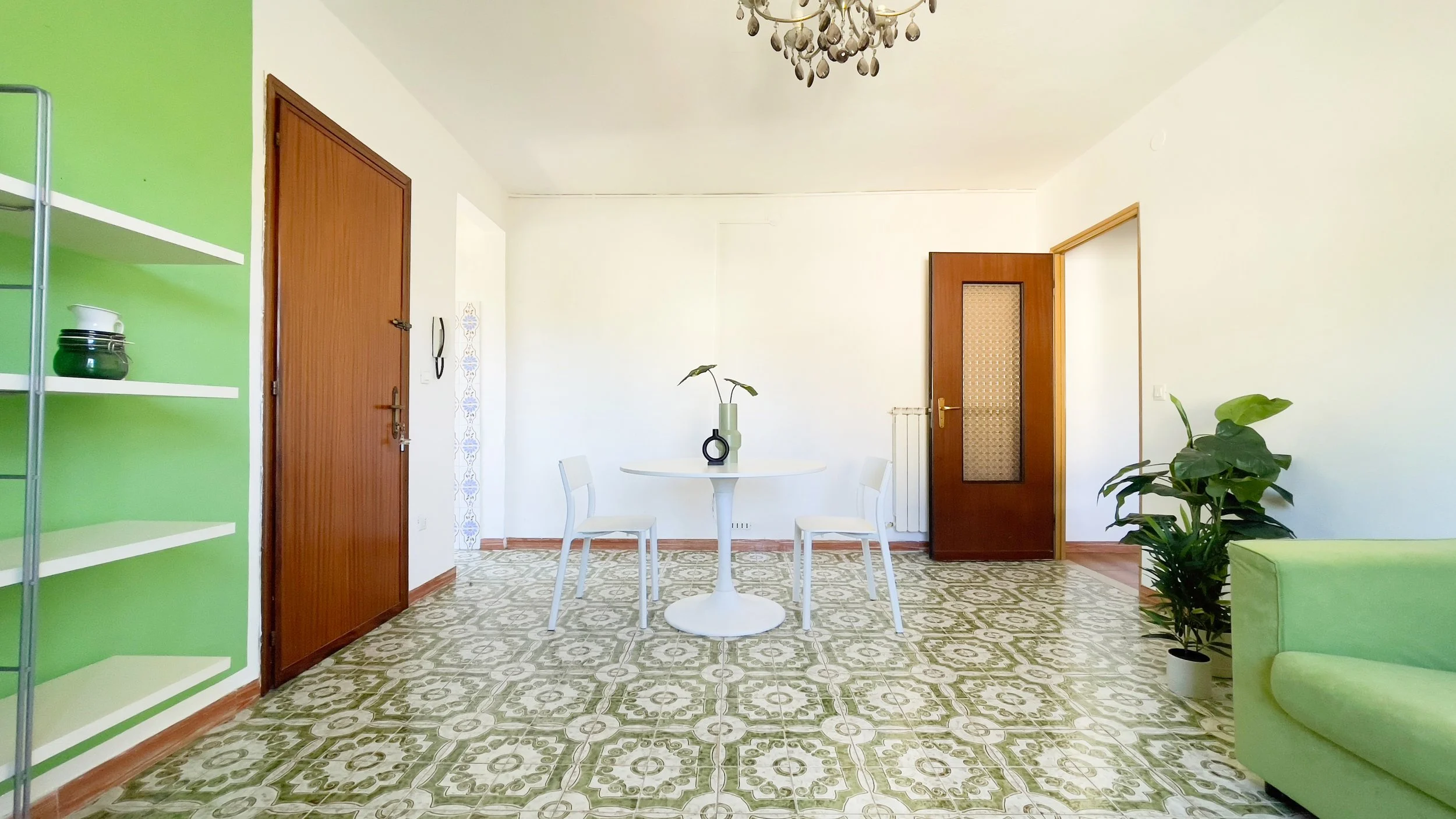 Stanza luminosa con pavimento decorato, pareti bianche, divano verde, tavolo rotondo bianco con due sedie, piante in vaso, e lampadario elegante.