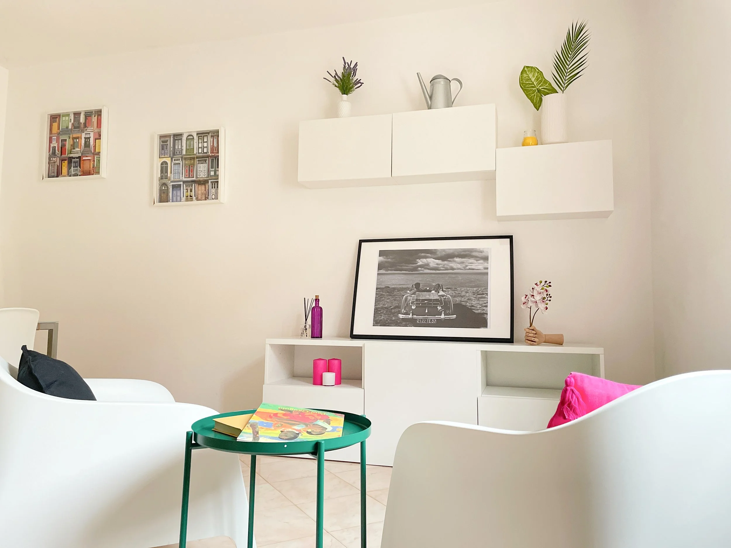Soggiorno moderno con mobili bianchi, tavolino verde, cuscini colorati, e decorazioni. Due quadri colorati e una foto in bianco e nero decorano la parete. Piante e candele completano l'arredo.