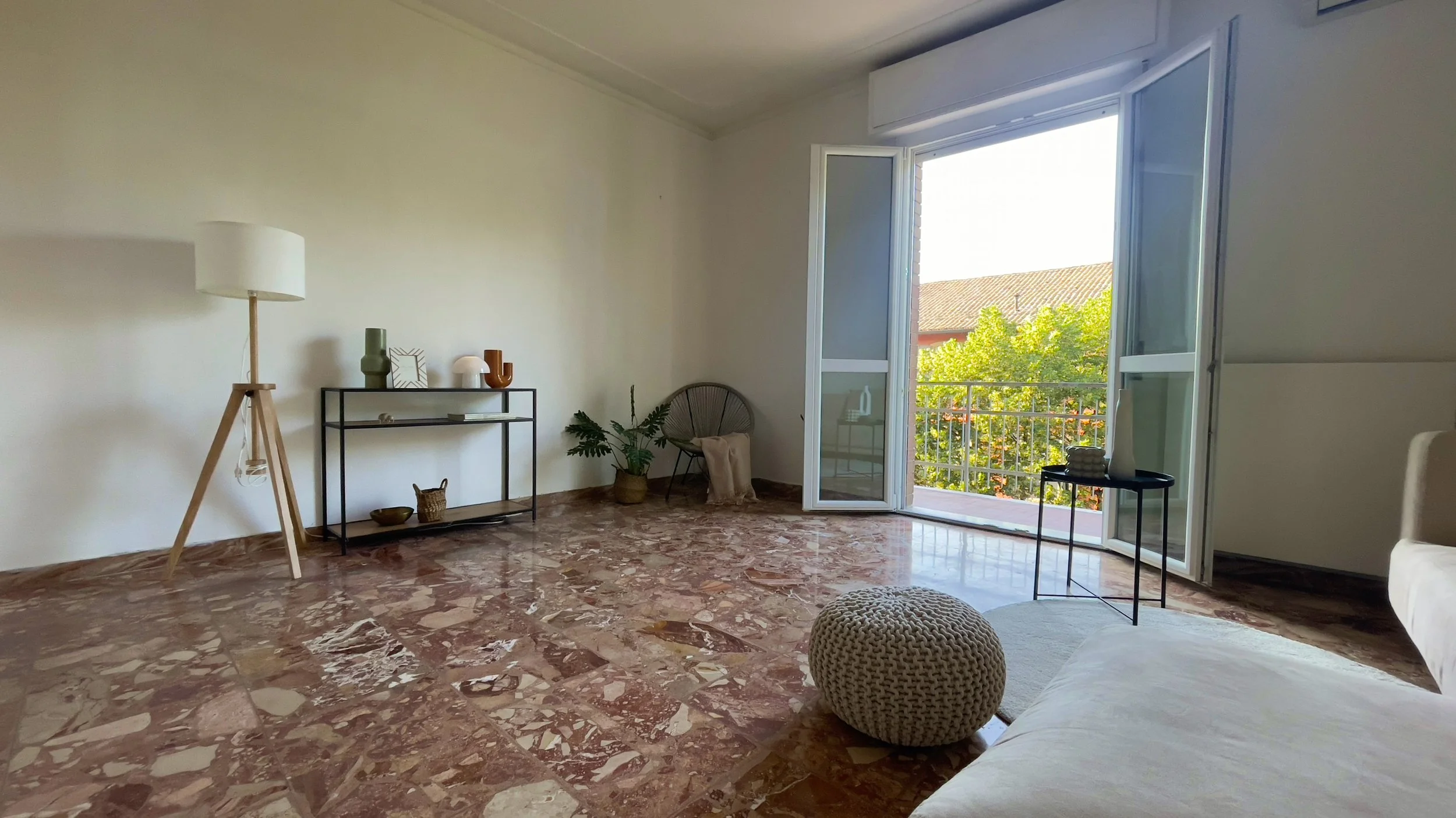 Sala con pavimento in marmo, lampada a stelo, mobile con oggetti decorativi, sedia con pianta, finestra aperta con vista sul balcone, pouf intrecciato sul pavimento.