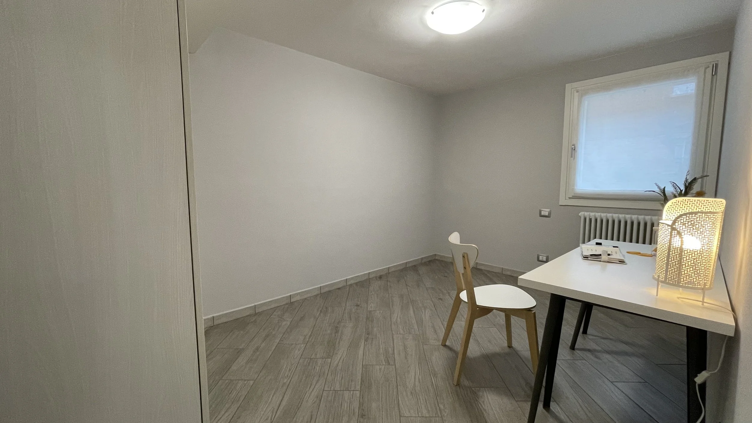 Interno di una stanza con scrivania bianca, sedia di legno, lampada accesa, finestra e pavimento in legno chiaro.