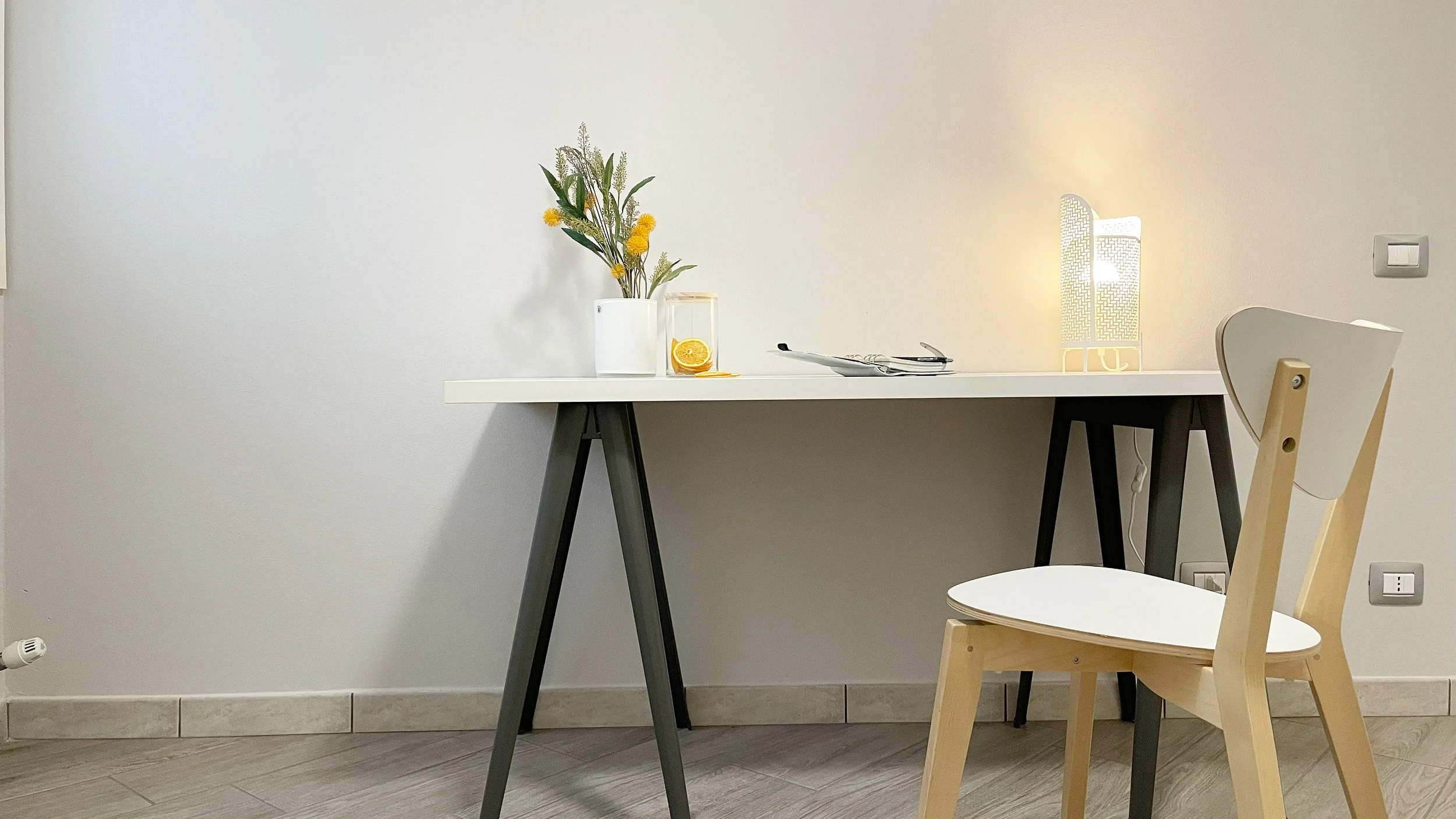 Scrivania moderna con sedia, lampada accesa, vasi di fiori gialli e tazza trasparente con fetta d'arancia. Pareti e pavimento chiari.
