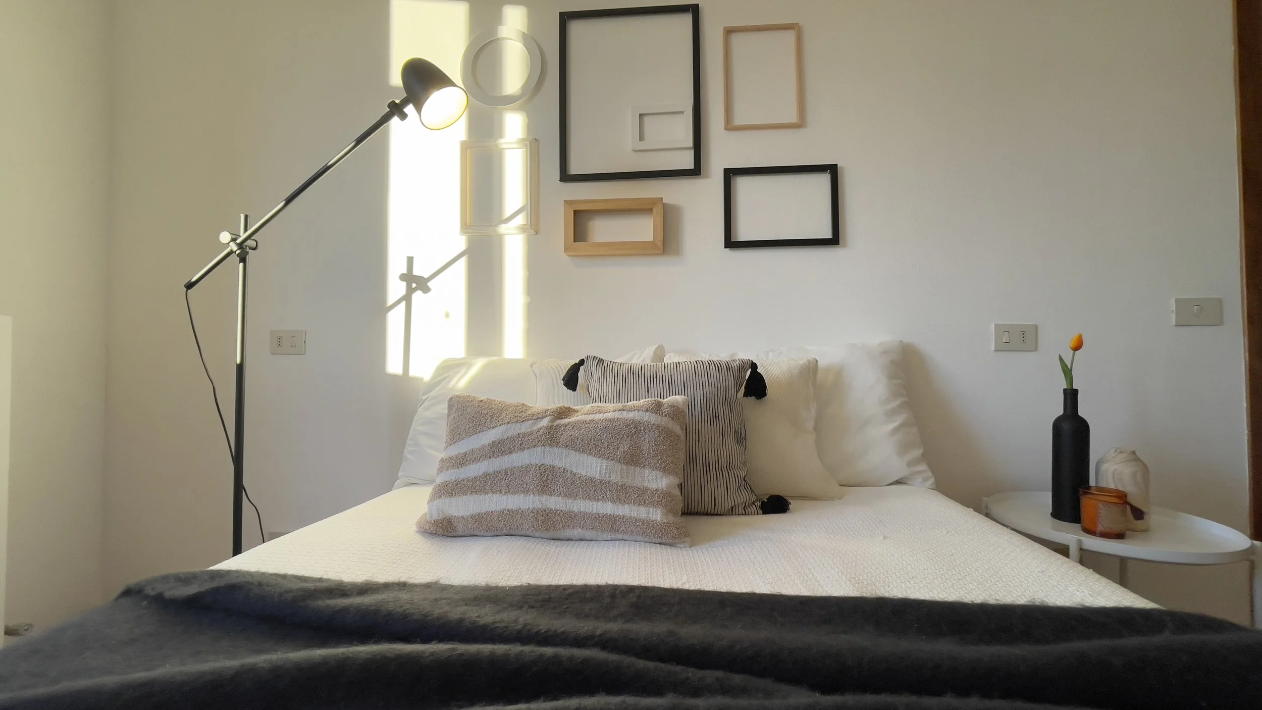 Camera da letto minimalista con letto bianco, cuscini decorativi, lampada da terra nera, cornici vuote appese al muro, tavolino con vaso nero e fiore, e candele.