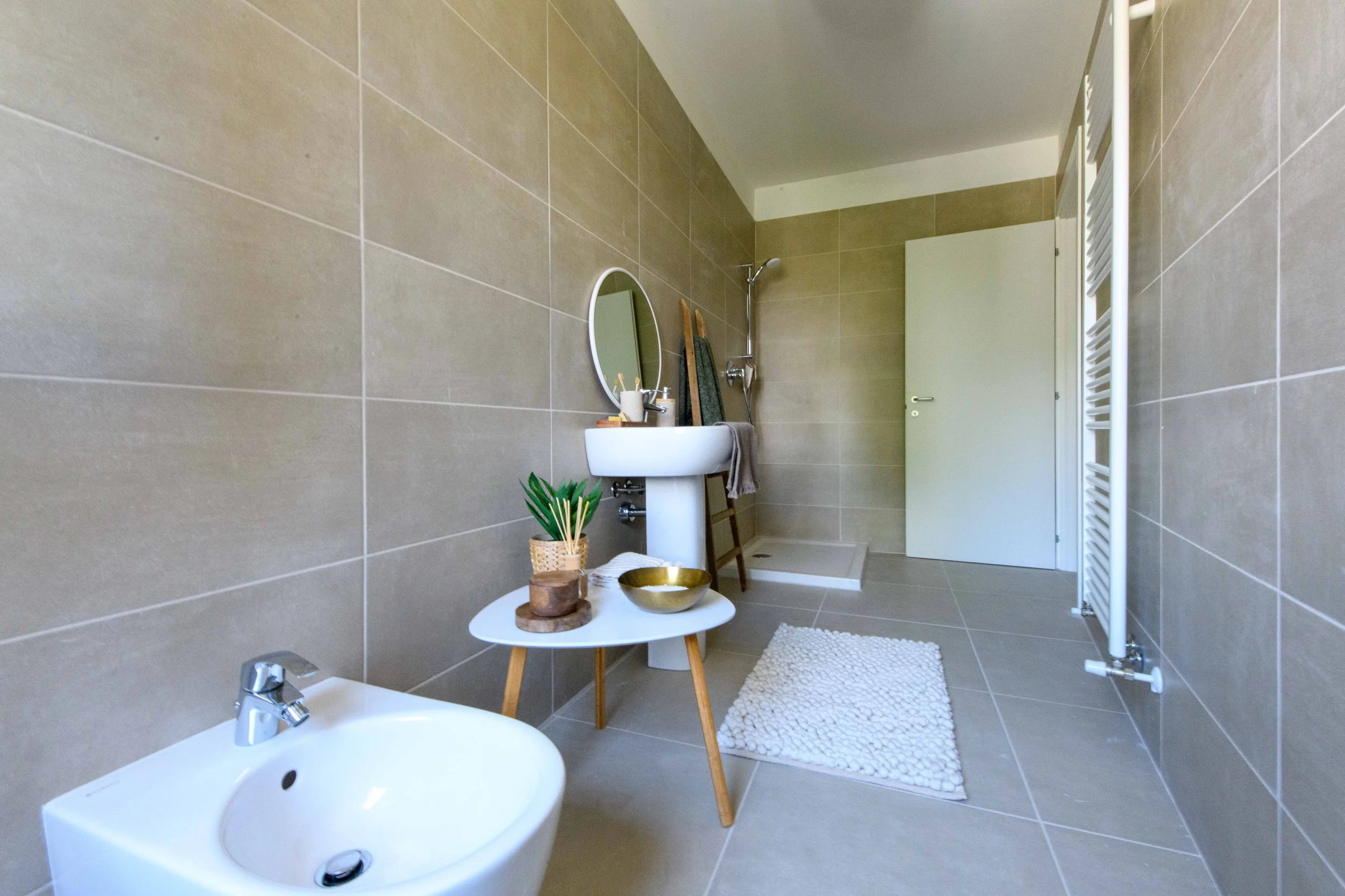 Bagno moderno con piastrelle color beige, lavandino, specchio rotondo, bidet, tavolino con decorazioni e doccia a vista.