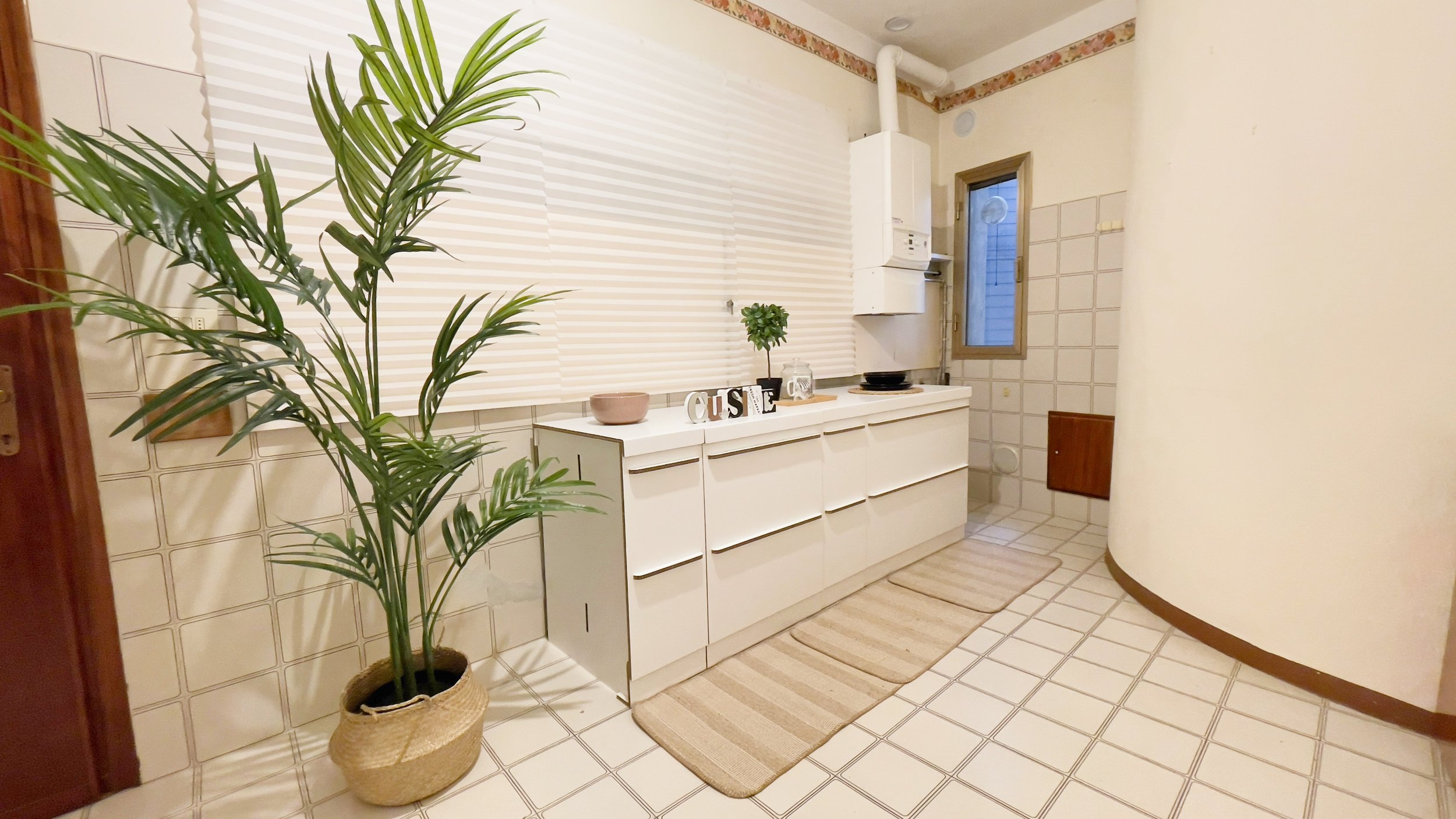 Cucina luminosa con mobili bianchi, pianta in vaso, tappeti beige, e decorazioni "CUISINE" su tavolo.