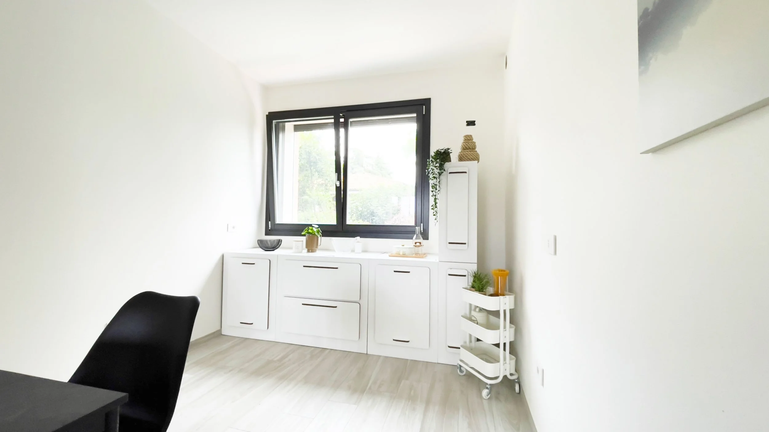 Interno di una cucina moderna con mobili bianchi, una sedia nera, un carrello bianco su ruote, e una finestra luminosa.