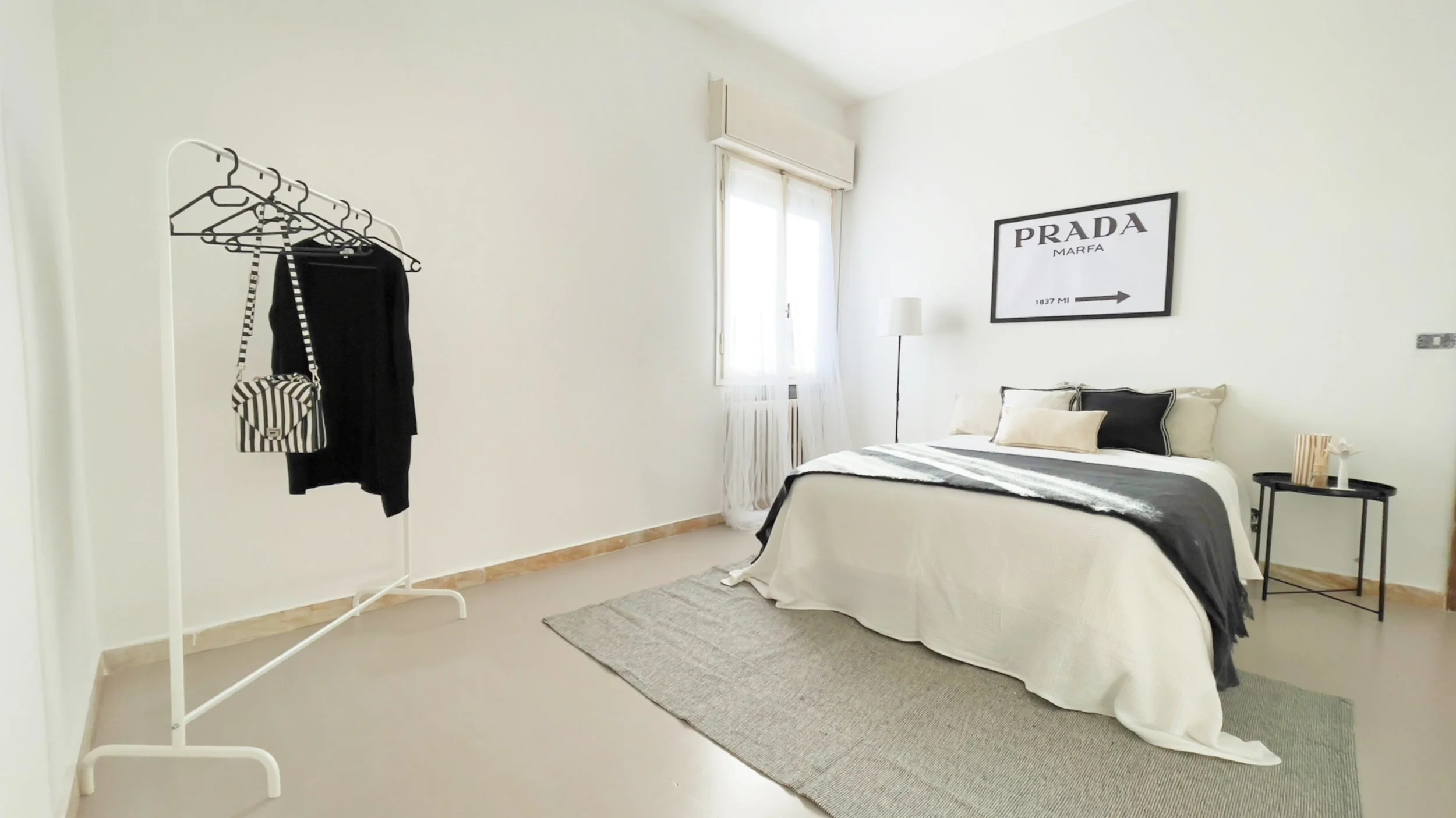 Camera da letto minimalista con letto matrimoniale, comodino, appendiabiti con vestiti e borsa, finestra con tende bianche e poster "Prada Marfa" sulla parete.
