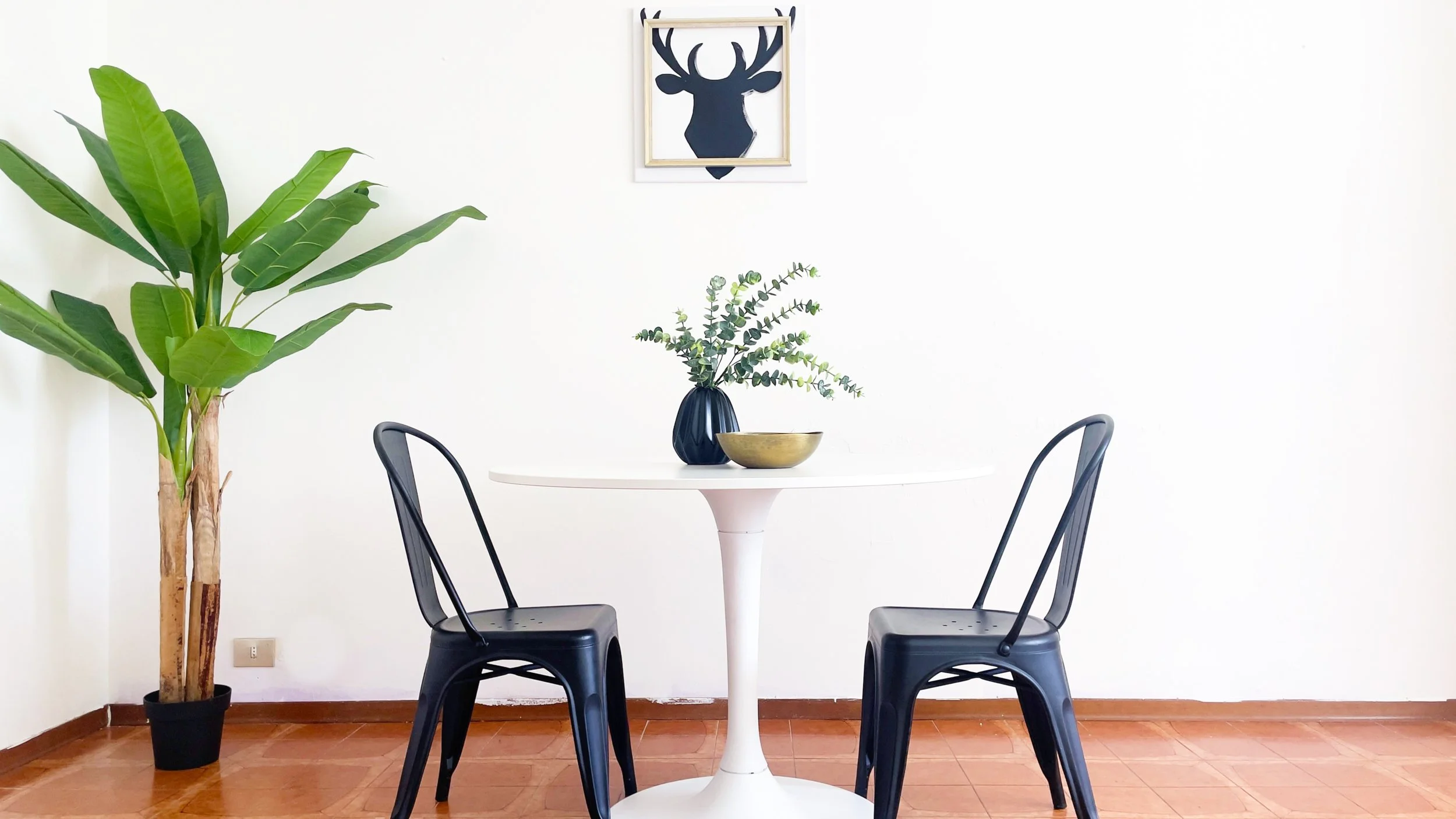 Sala da pranzo moderna con tavolo bianco, due sedie nere, pianta verde e quadro di testa di cervo.
