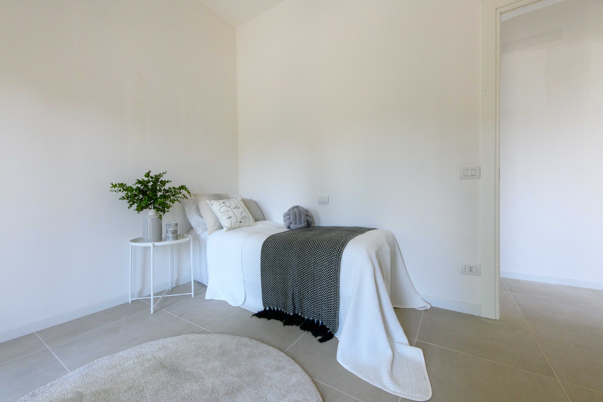 Camera da letto minimalista con letto singolo, copriletto bianco e coperta grigia, cuscini decorativi e una pianta su un tavolino bianco.