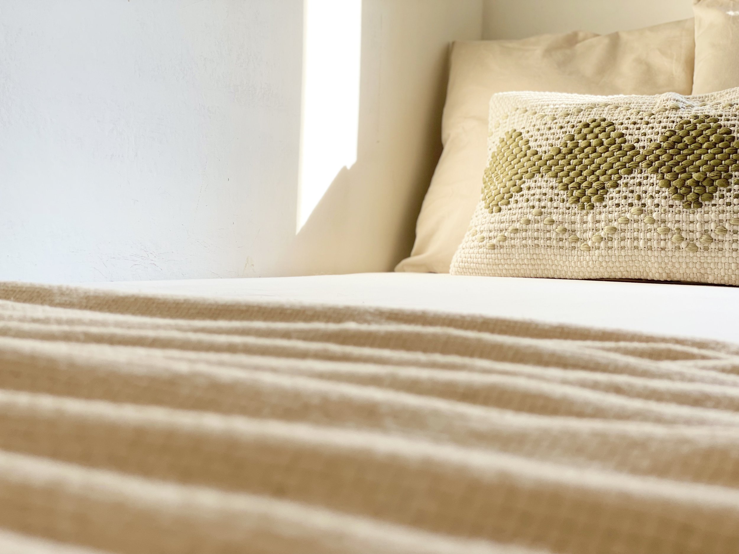 Letto con copriletto beige e cuscino decorativo a maglia su una parete bianca illuminata dal sole.