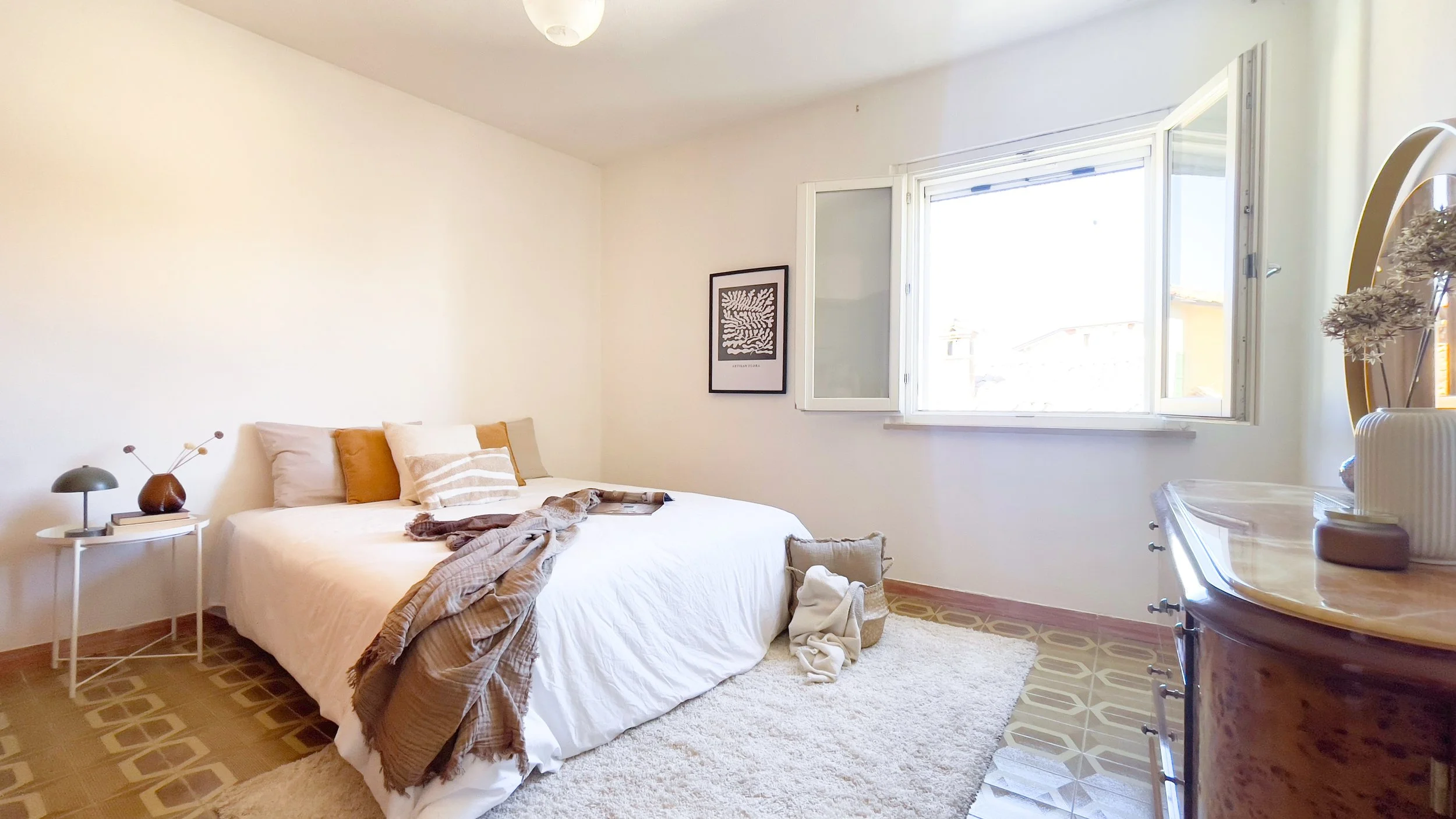 Camera da letto luminosa con un letto matrimoniale, cuscini e una coperta. Una finestra aperta lascia entrare la luce naturale. Tavolino con lampada e decorazioni a destra del letto. Un mobile con piante e uno specchio rotondo sulla destra.