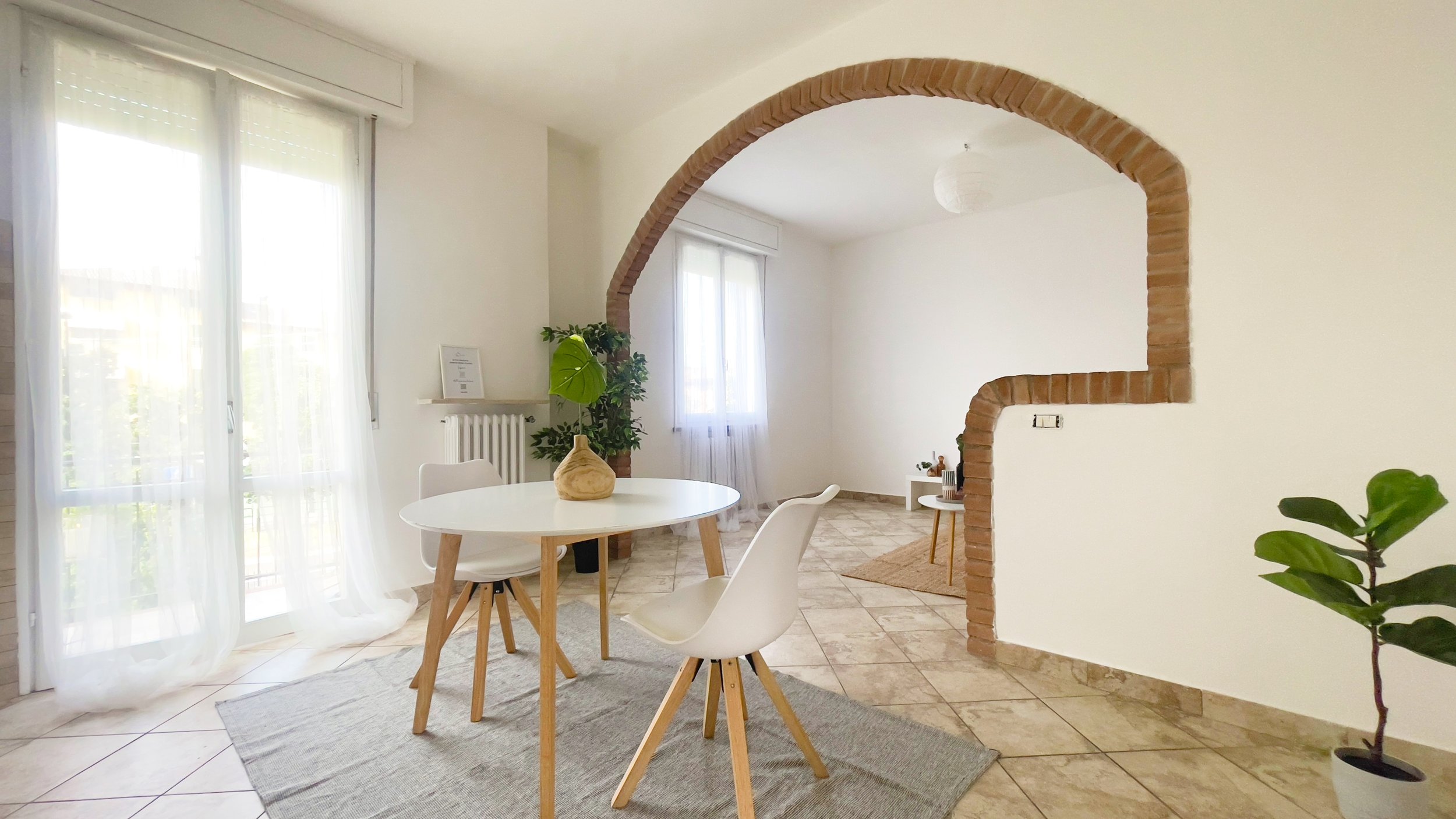 Sala da pranzo moderna con tavolo rotondo bianco, sedie bianche, pareti bianche, arco in mattoni, pavimento piastrellato e piante decorative.