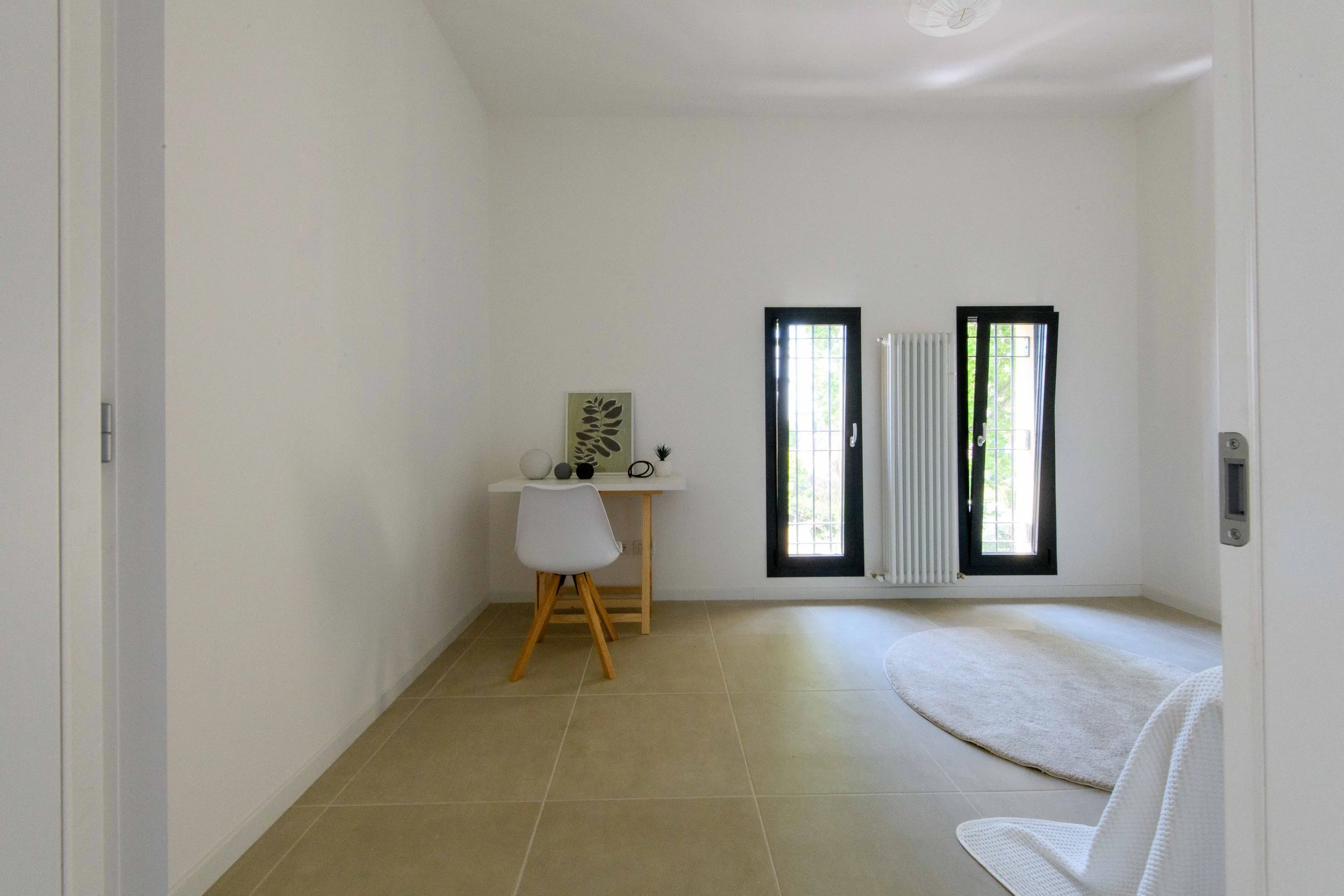 Stanza minimalista con scrivania bianca, sedia bianca, due finestre con telai neri, radiatore bianco e tappeto circolare chiaro.