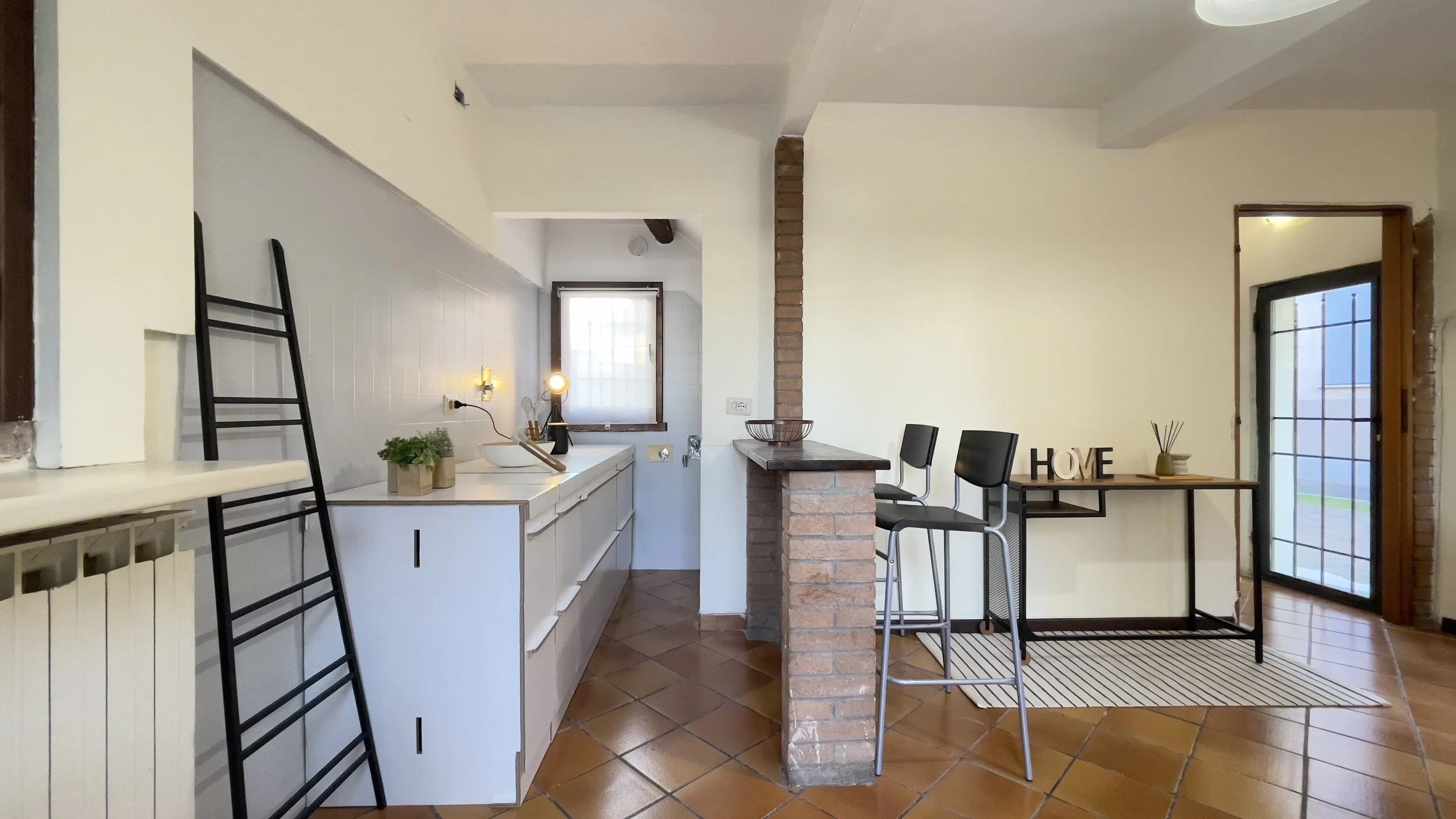 Interno di una cucina moderna con pavimento in piastrelle marroni, mobili bianchi, e tavolo con sgabelli alti. Accanto c'è una piccola area con decorazioni e la parola 'HOME'. Una porta finestra conduce all'esterno.
