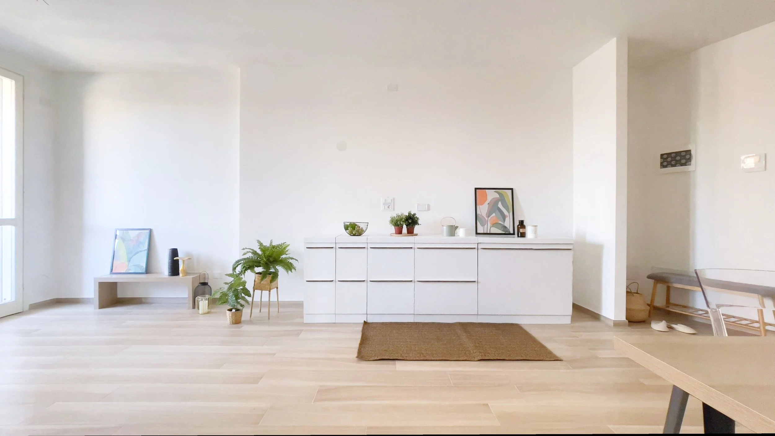 Soggiorno minimalista con mobili bianchi, piante, tavolo in legno chiaro, decorazioni artistiche e luce naturale.