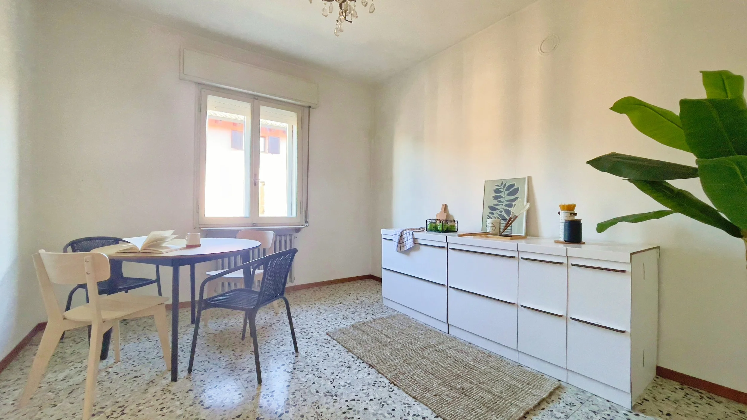 Interno di una stanza luminosa con un tavolo circolare e sedie, una finestra a sinistra, mobili bianchi sulla destra, una pianta verde e un tappeto sul pavimento.