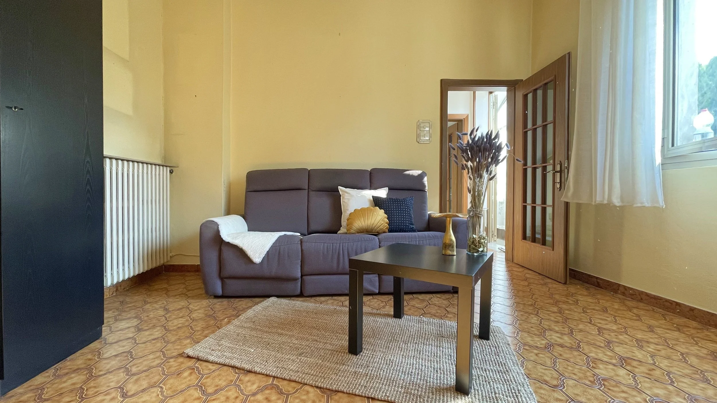 Soggiorno con divano grigio e cuscini, tavolino nero con decorazione floreale, tappeto beige, armadio nero e porta con vetri; pavimento in piastrelle marroni e pareti gialle.
