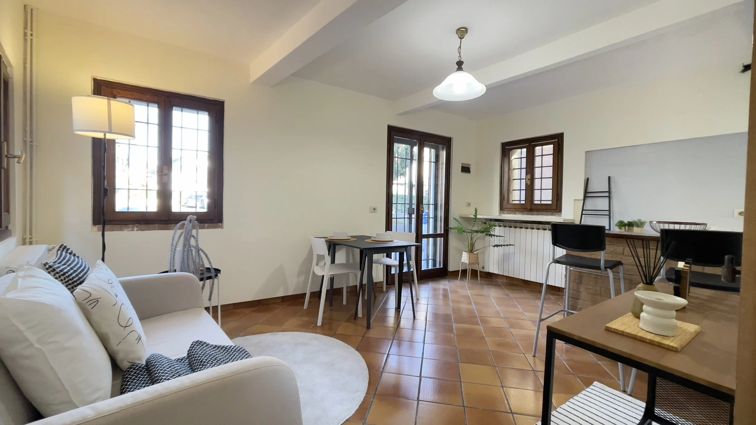 Interno di un soggiorno moderno con pavimento in ceramica marrone, pareti bianche e soffitto con una lampada centrale. A sinistra, un divano bianco con cuscini decorativi. Al centro, un tavolo con due sedie. A destra, una finestra grande e una scrivania con accessori. Pianta decorativa vicino al radiatore.