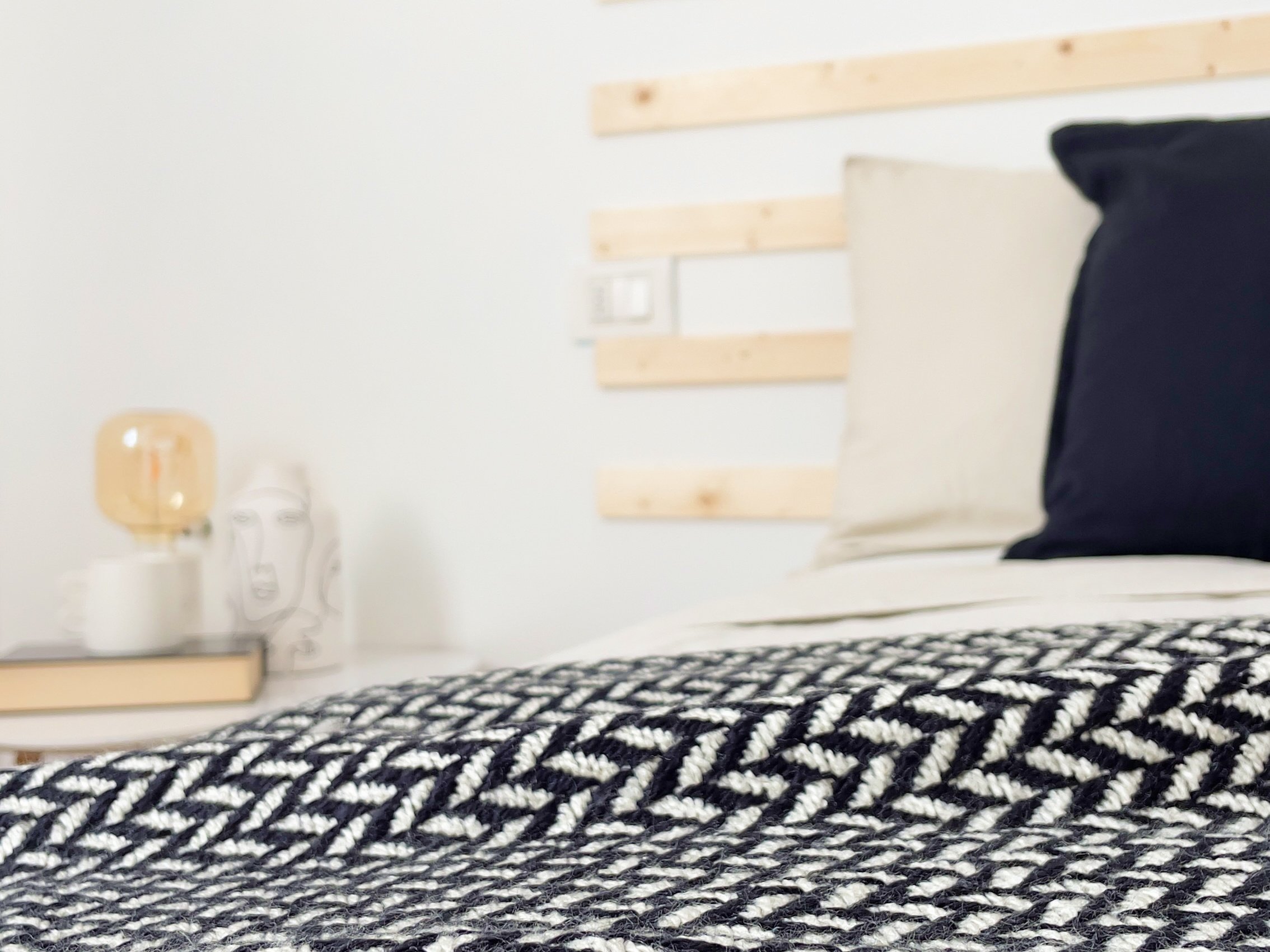 Interno di una camera moderna con letto sfatto, cuscini neri e beige, coperta a motivi geometrici, piccolo tavolo con lampada e libro.