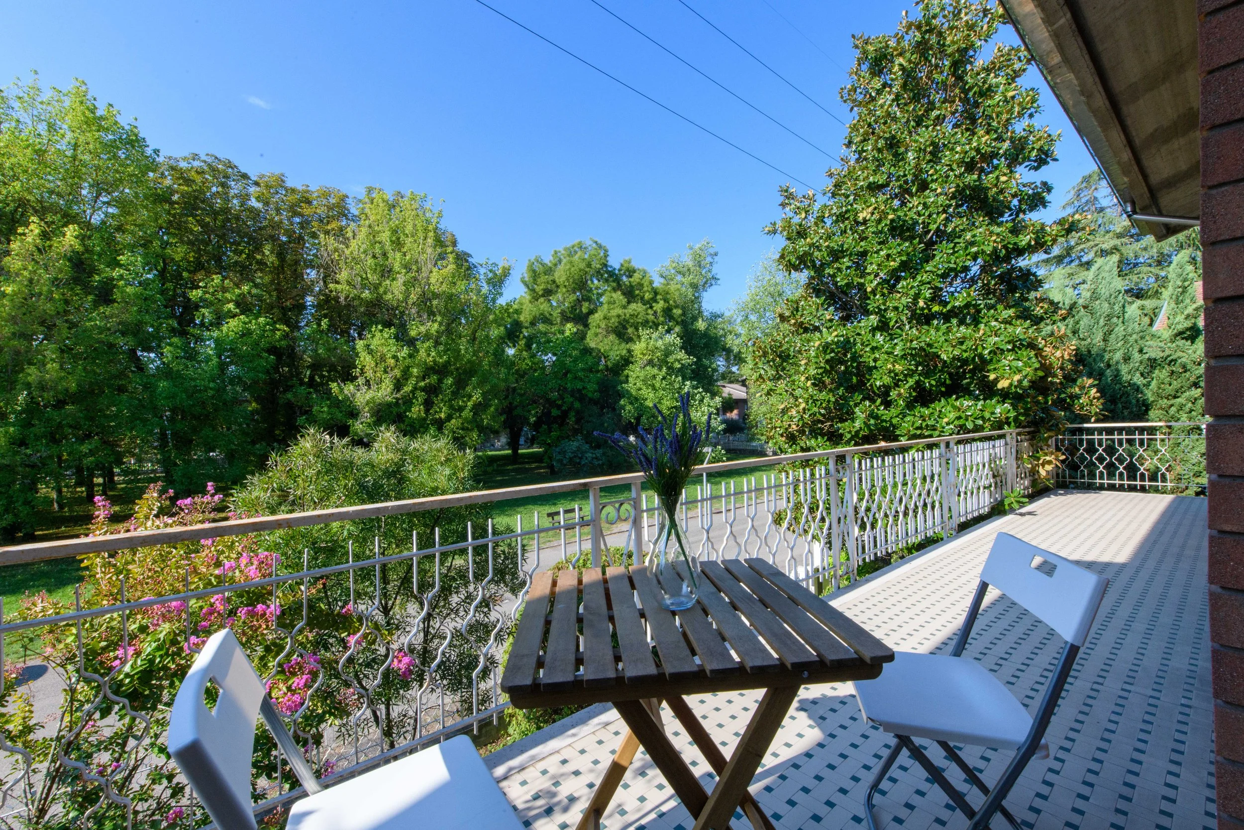 Terrazza con tavolo e sedie, circondata da alberi e vegetazione. Cielo soleggiato e limpido.
