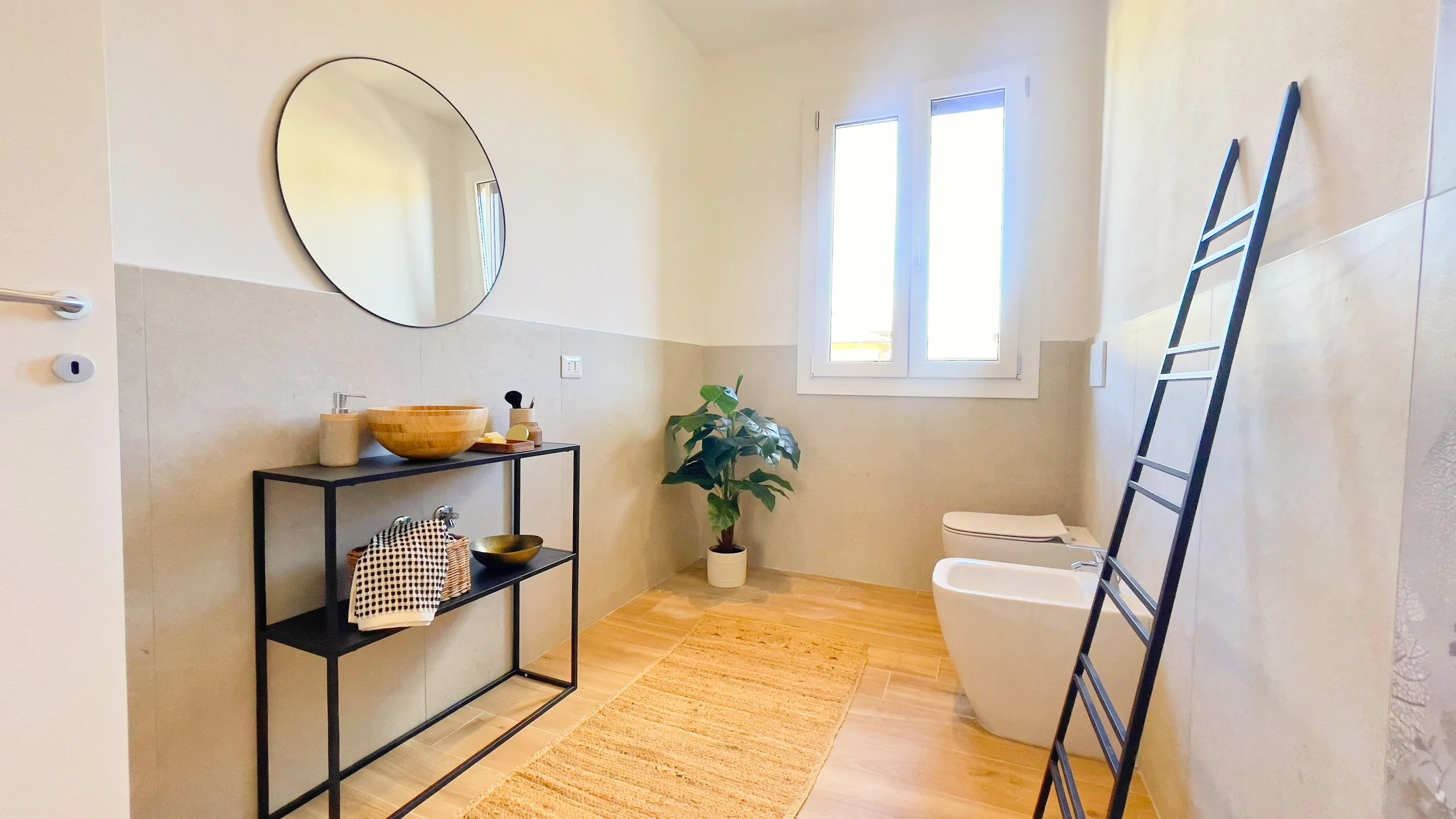 Bagno moderno con specchio rotondo, lavandino con ciotola di legno, pianta in vaso, finestra, bidet, WC e scala porta asciugamani.