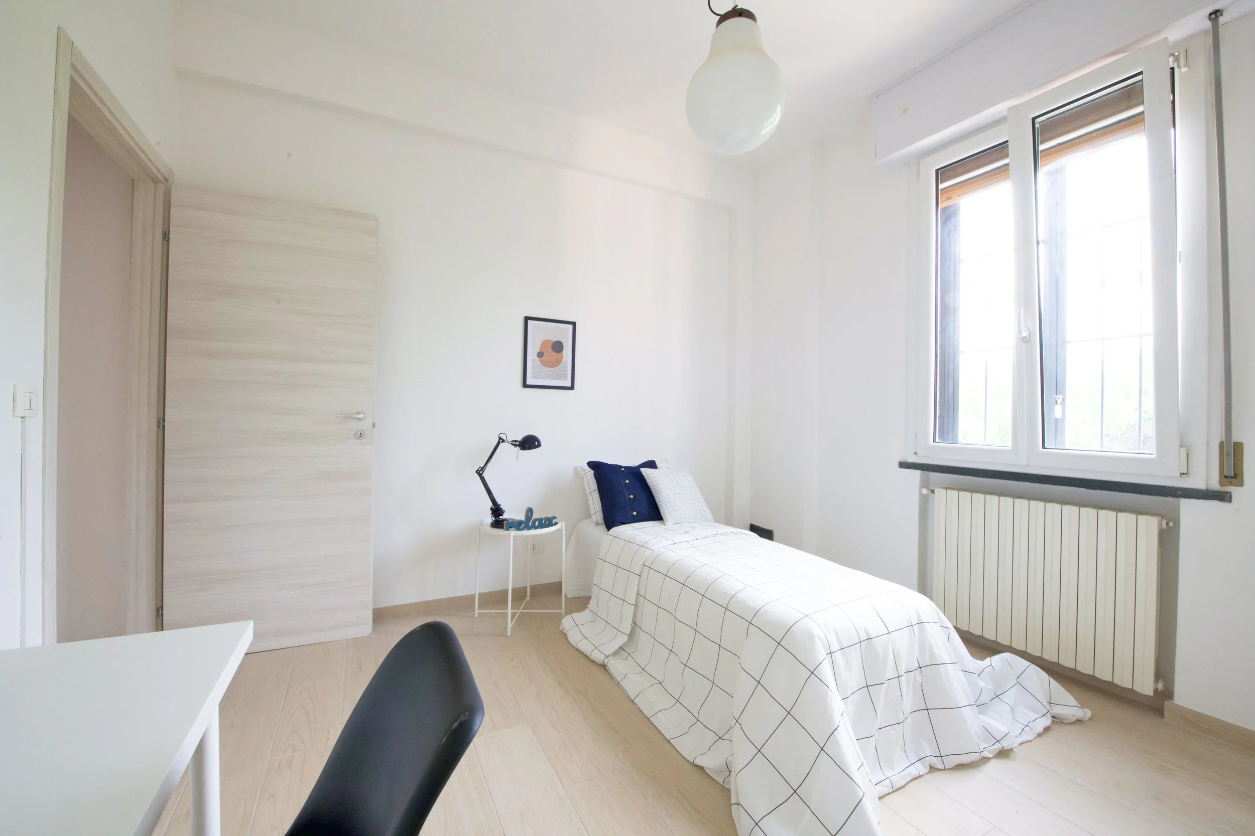 Camera da letto minimalista con letto singolo, coperta a quadri, scrivania bianca con sedia nera, lampada da tavolo, finestra luminosa, e decorazioni semplici.