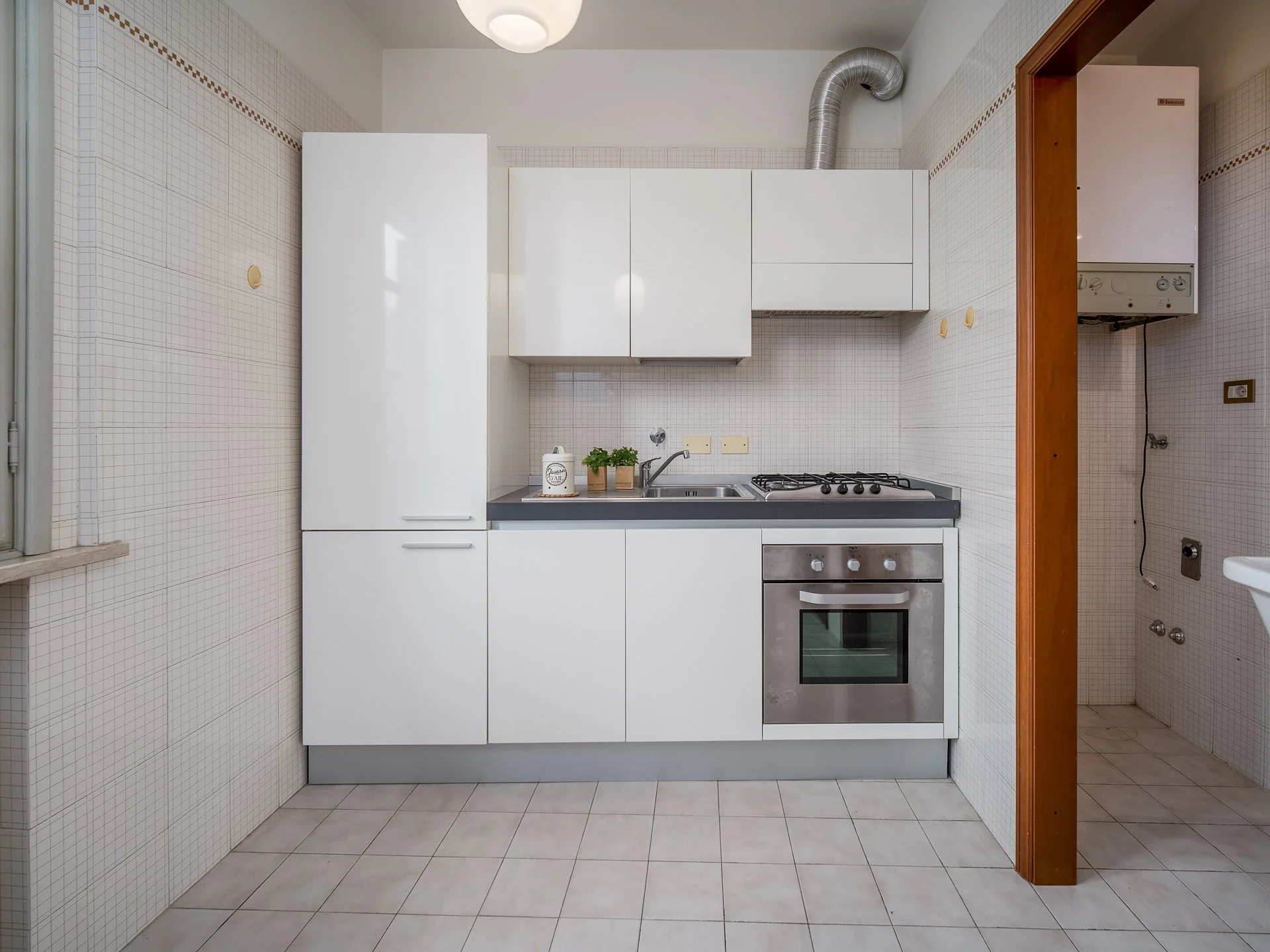 Cucina moderna con mobili bianchi, forno, piano cottura, lavello e cappa in un ambiente piastrellato.