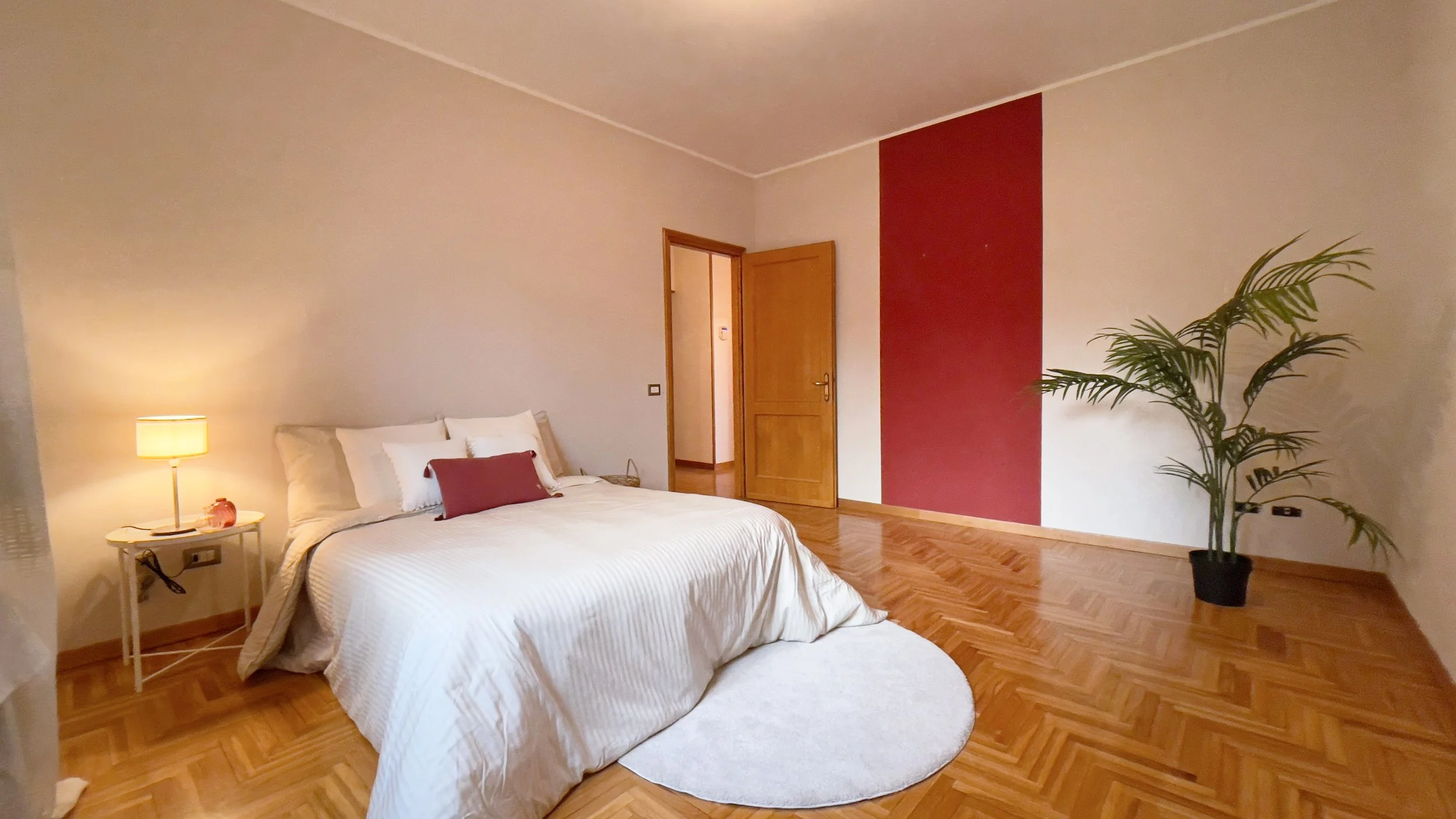 Camera da letto con letto matrimoniale, lampada su un comodino, parete bicolore rosso e bianco, pianta ornamentale, pavimento in parquet.