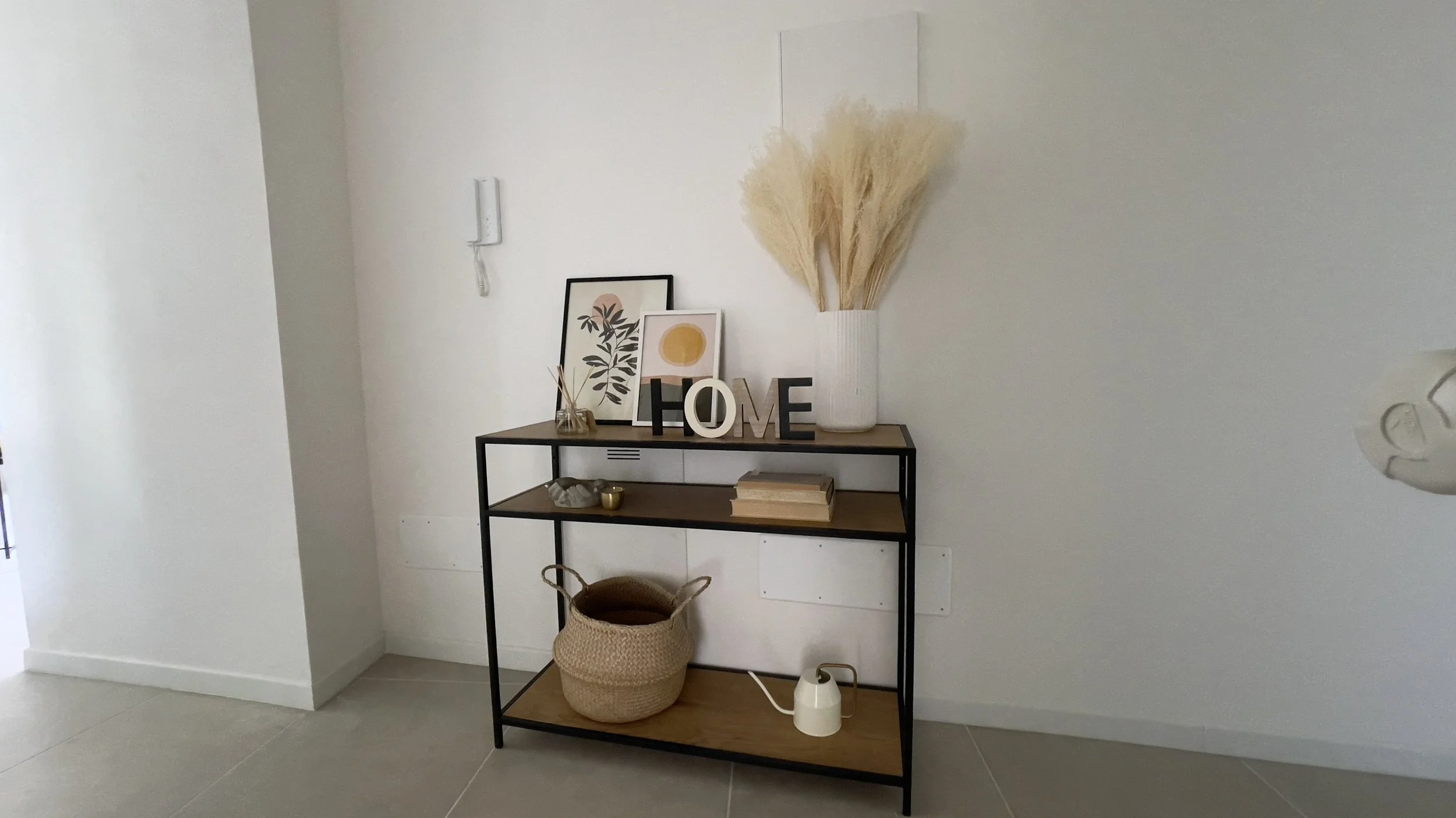 Scaffale con decorazioni, piante secche in vaso bianco, cornici con stampe, lettere "HOME", diffusore di fragranze, cesti di vimini e annaffiatoio.